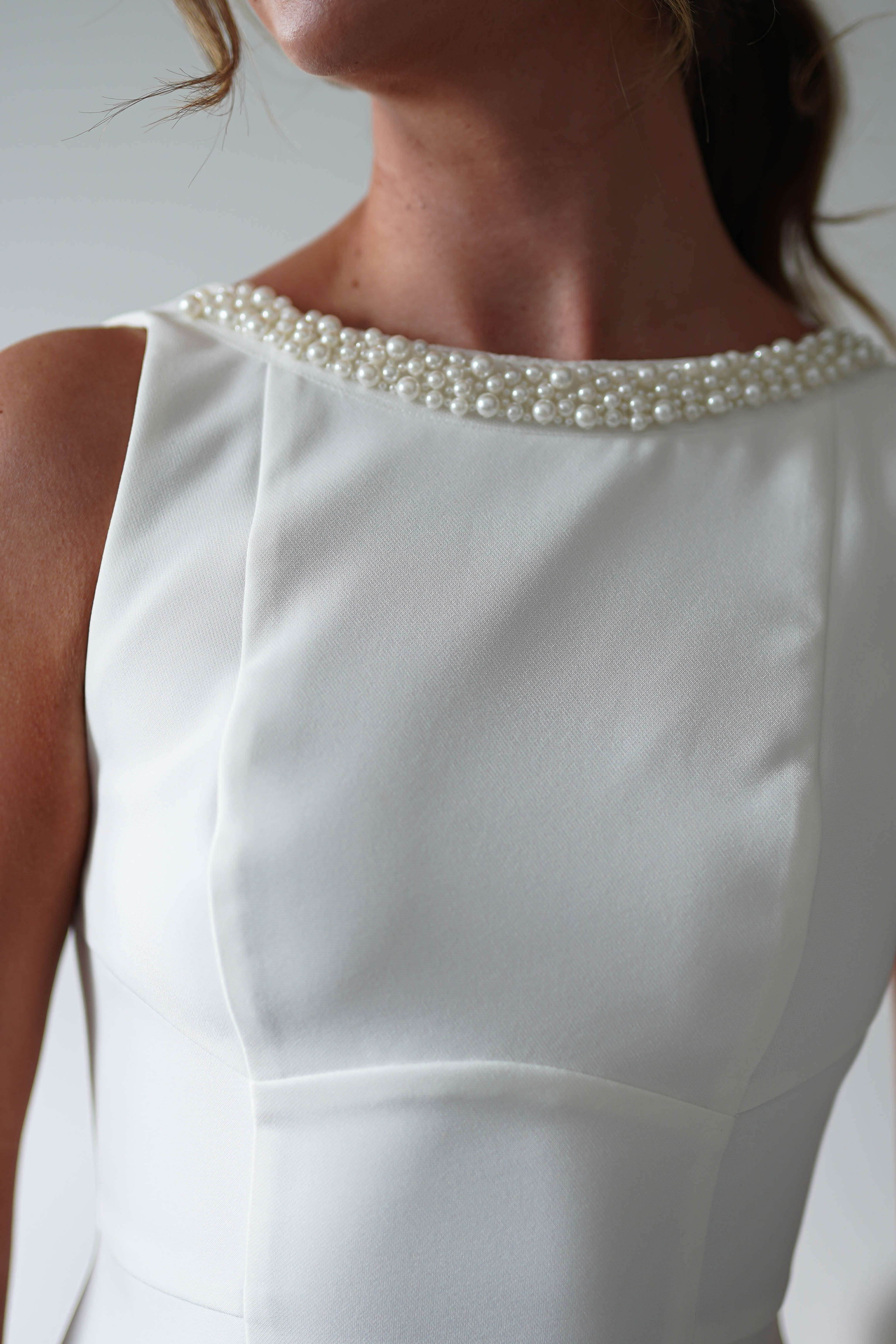 A close-up of a woman wearing a sleeveless white mini dress with a fitted bodice and a round neckline adorned with pearl detail—perfect for parties. Her face is partially out of frame, and her brown hair is tied back.