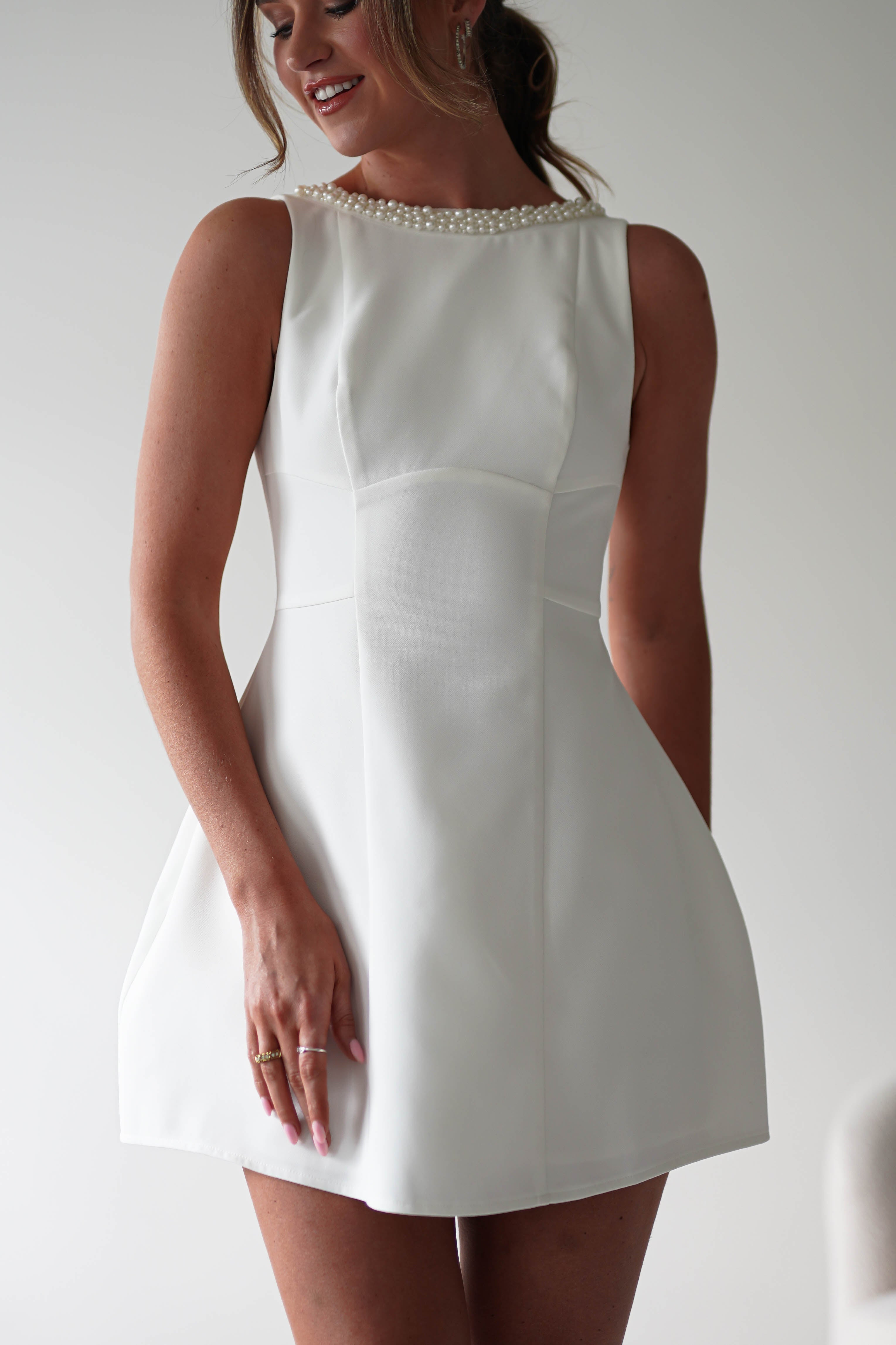 A woman wearing a sleeveless, white mini dress with pearl detail around the neckline, smiling and posing with one hand on her hip—perfect for parties.