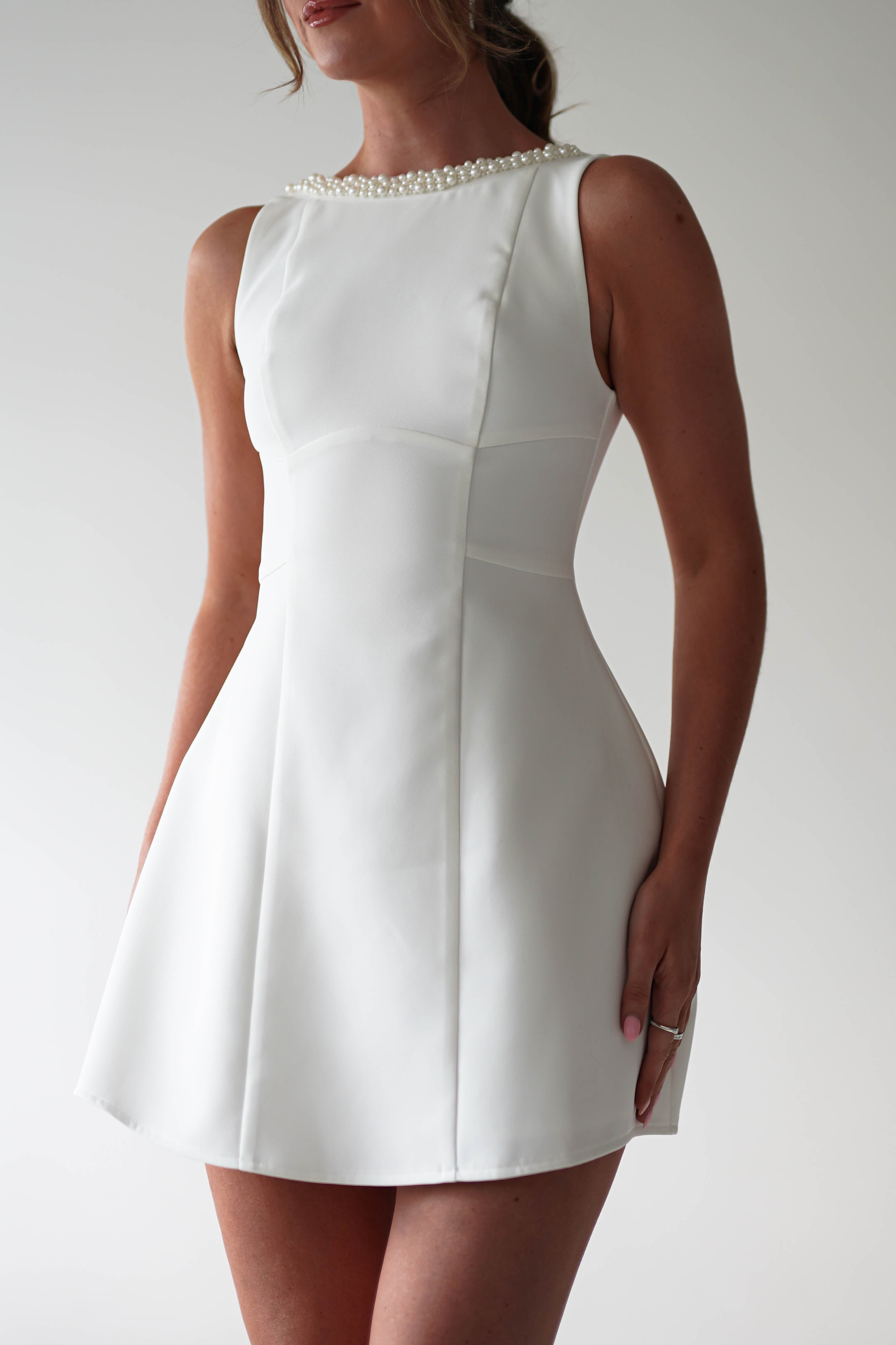 A woman wearing a sleeveless, fitted white mini dress with a flared skirt and pearl detail along the neckline stands against a plain light background—perfect for parties.