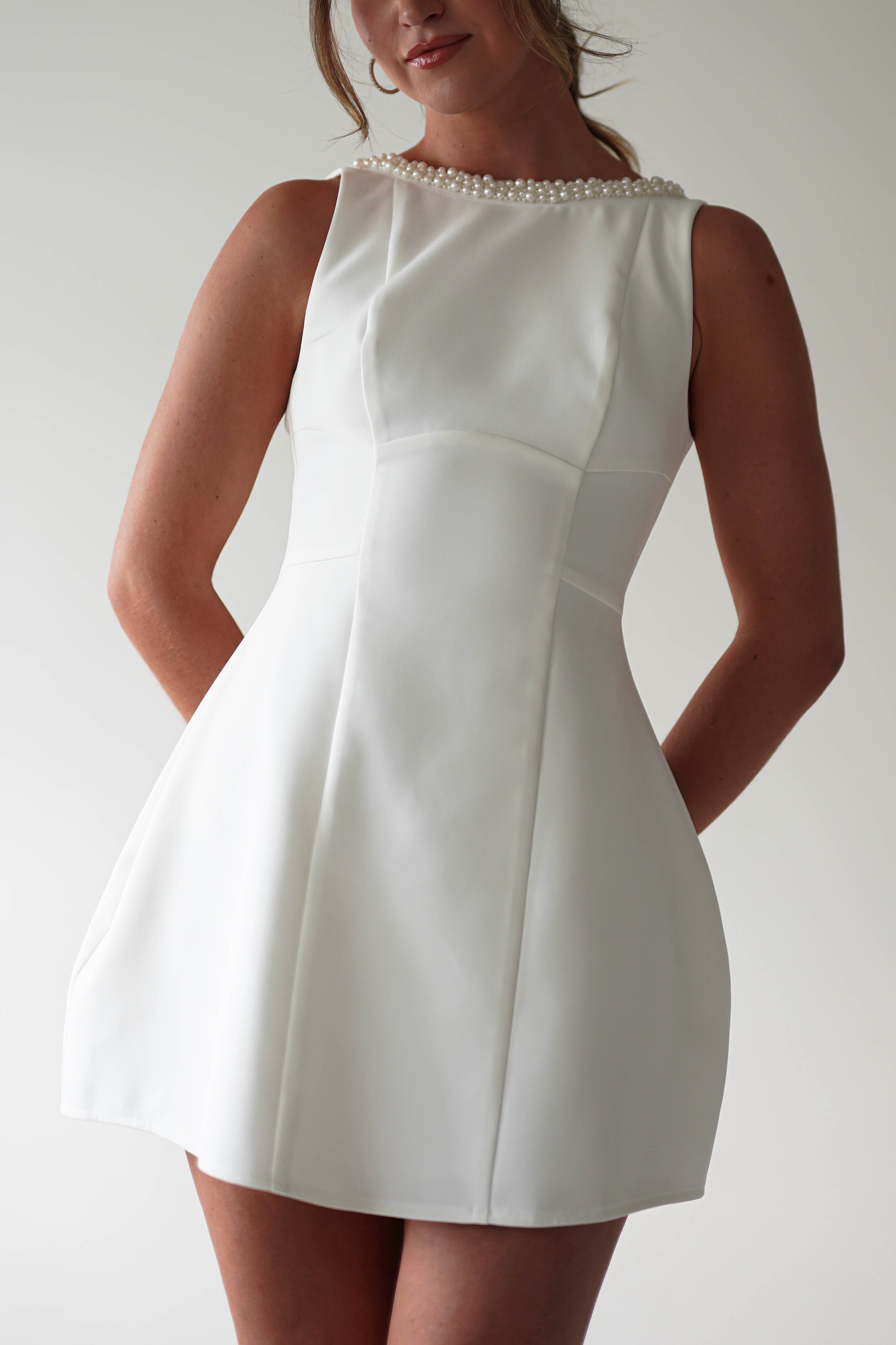 A woman in a sleeveless, fitted white mini dress with pearl detail at the neckline stands with her hands behind her back against a plain light background—an elegant choice for parties.