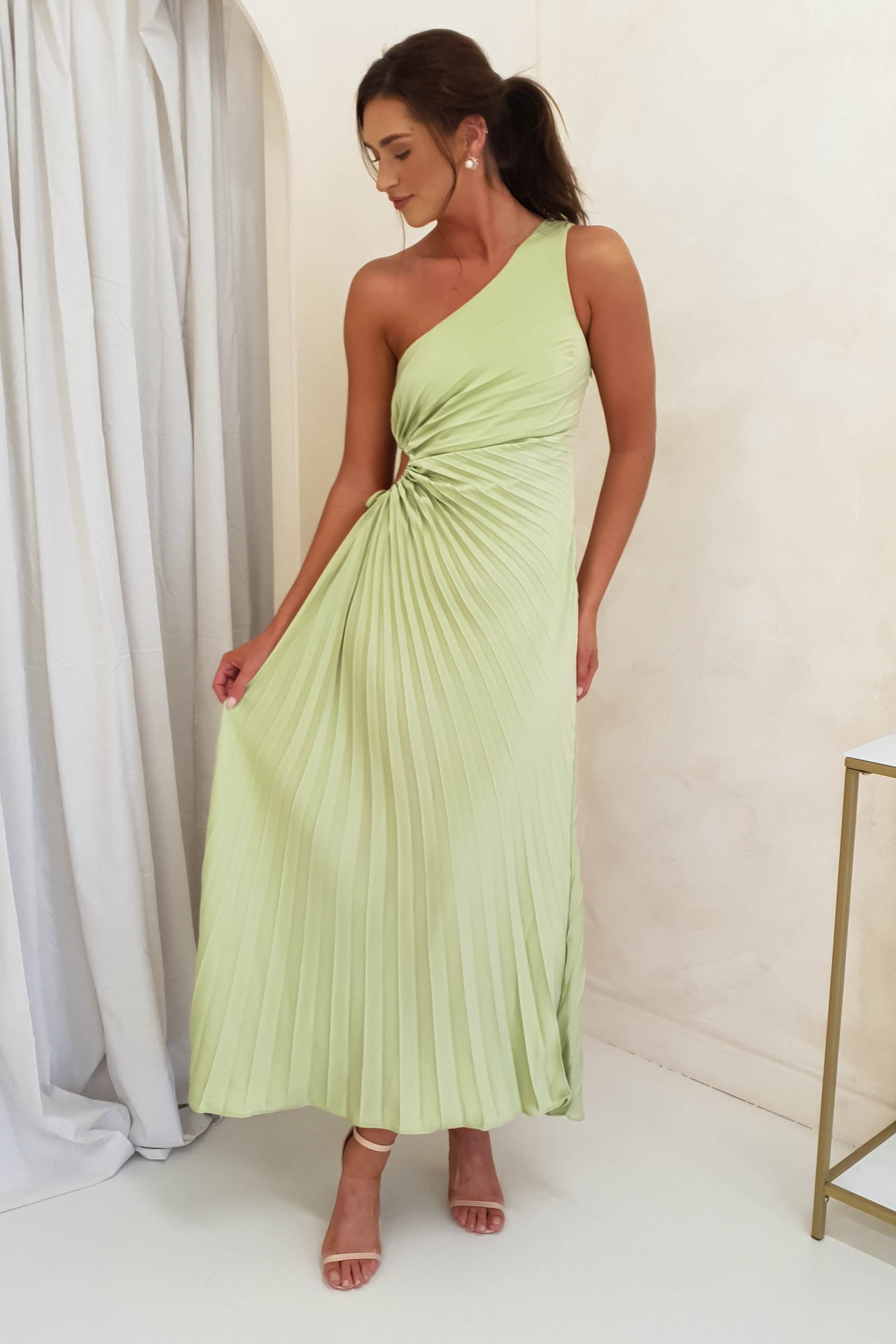 A woman in a light green, one-shoulder pleated maxi dress stands indoors, holding her skirt out to the side. Her dark hair is tied back and she wears nude heels—perfect formal events attire against simple white curtains.