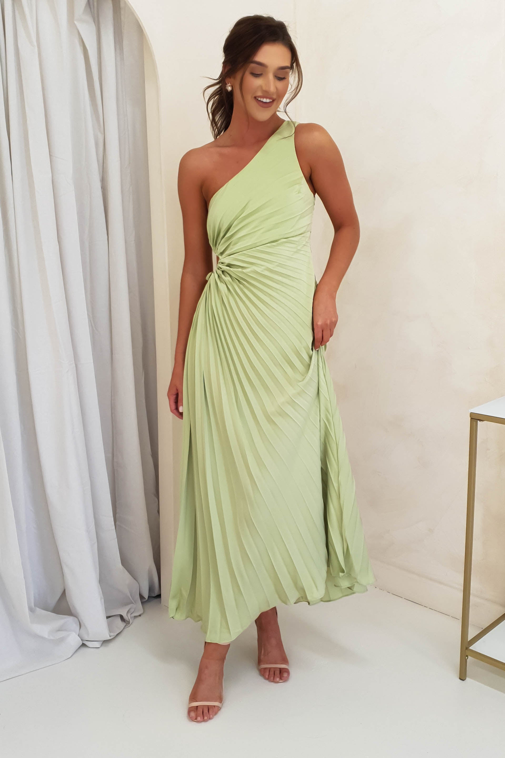 A woman wearing a light green, one-shoulder pleated maxi dress stands indoors on a white floor, smiling with one hand by her side and the other touching the dress. She wears nude heels and has her hair styled in loose waves—perfect formal events attire.