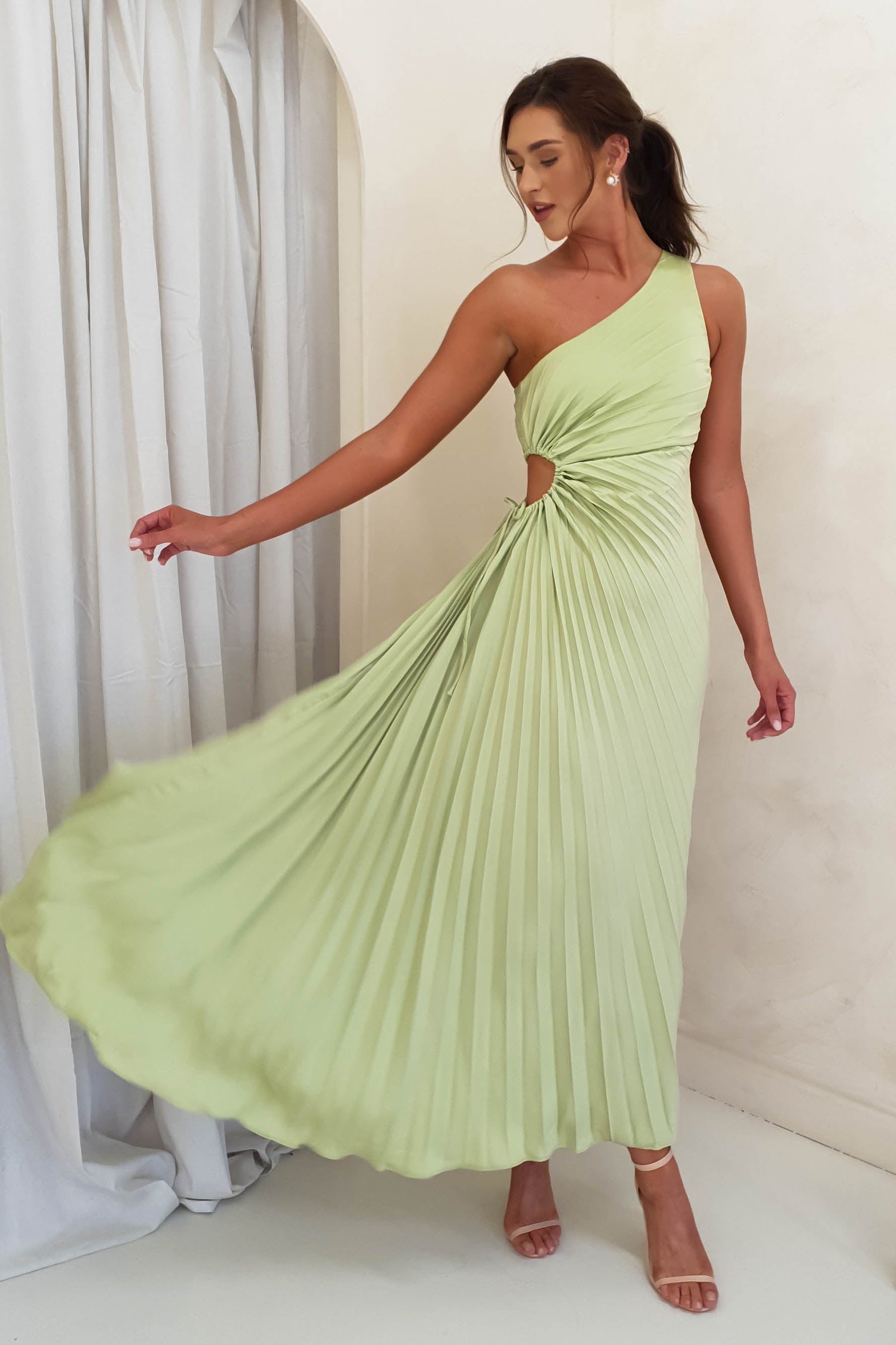 A woman wearing a light green, one-shoulder maxi dress with pleats and a side cut-out poses gracefully, holding out her skirt. Perfect for formal events attire, she stands indoors against a cream-colored wall and white curtains, in strappy heels.
