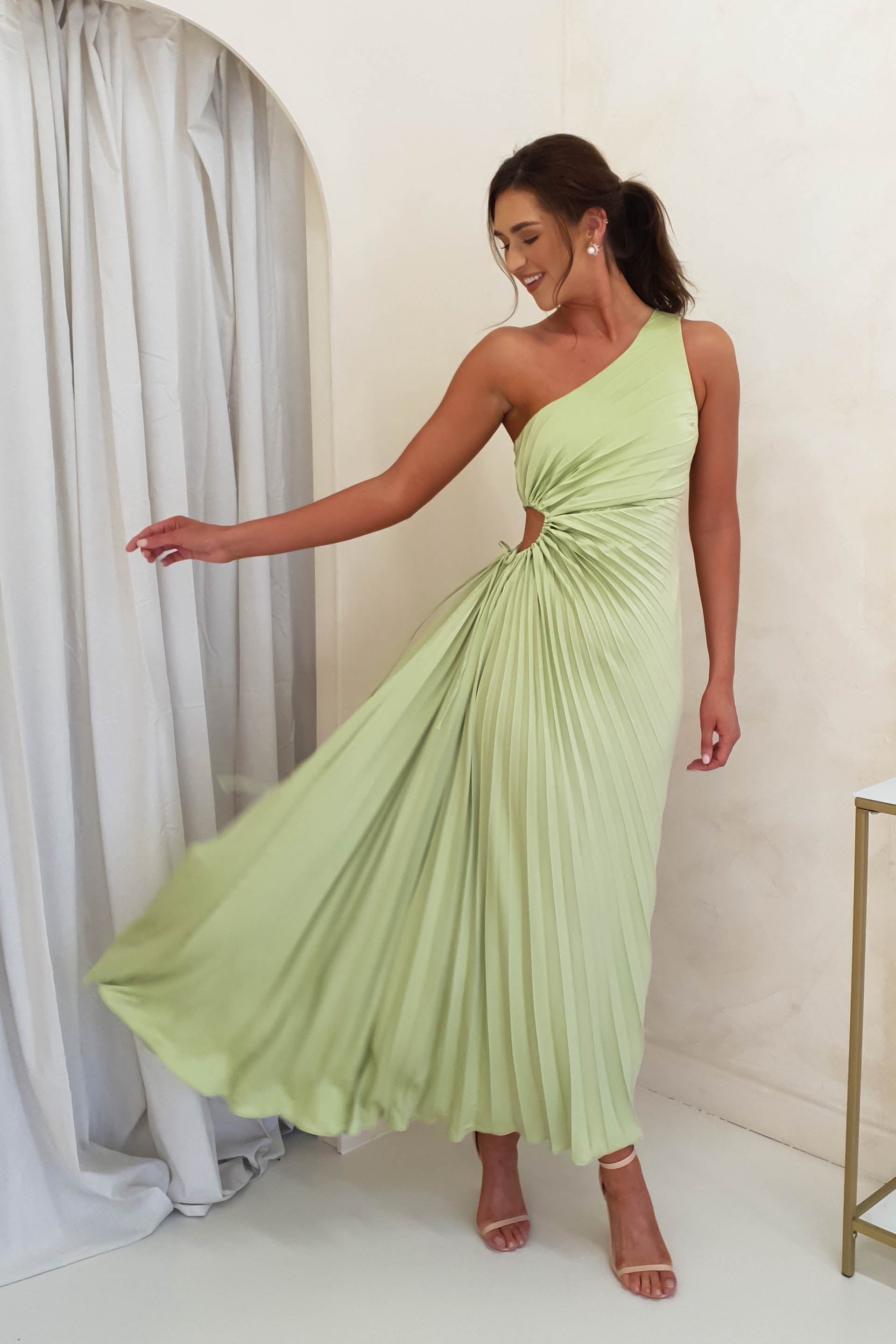 A woman wearing a light green, one-shoulder maxi dress poses gracefully, holding out her pleated skirt. She stands indoors near white curtains, smiling and looking down, with one arm extended and her hair styled in a ponytail.
