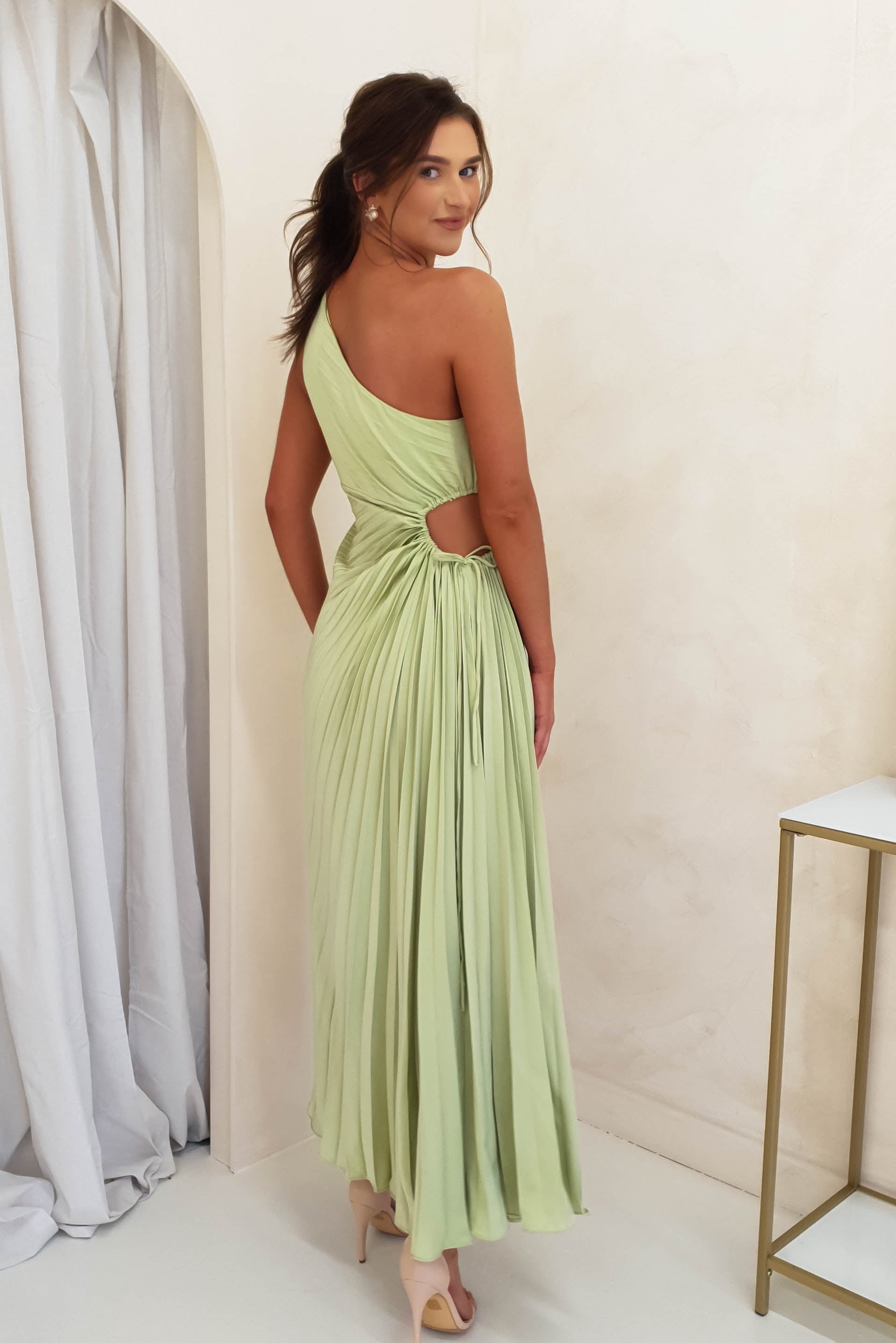 A woman in a light green, one-shoulder pleated maxi dress with a waist cut-out stands in a minimal room, looking over her shoulder. Ideal for wedding guests or formal events, she pairs her look with nude heels and a sleek ponytail.