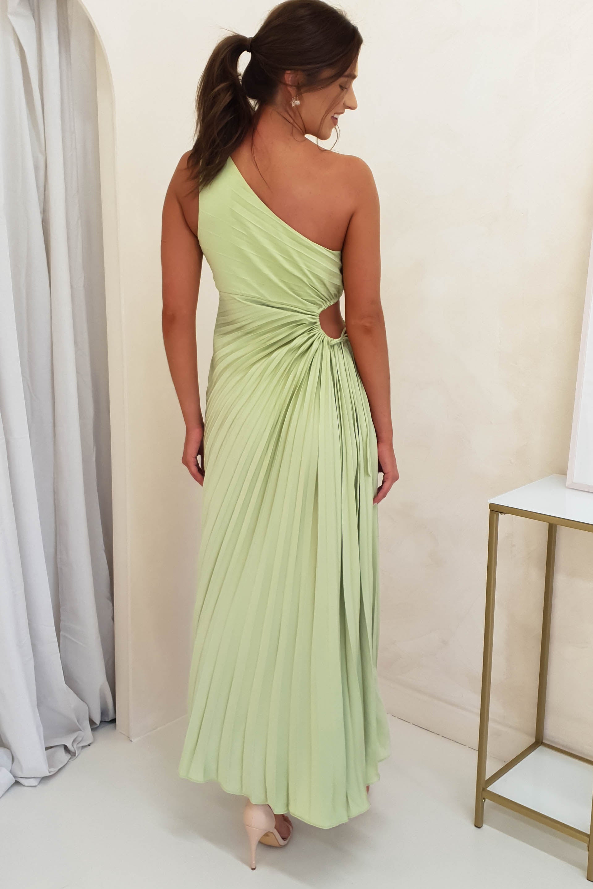 A woman in a pleated maxi dress with a one-shoulder design and side cut-out stands facing away. Her hair is in a ponytail and she wears nude high heels, standing on a light floor by a table and curtain—an elegant choice for formal events attire.