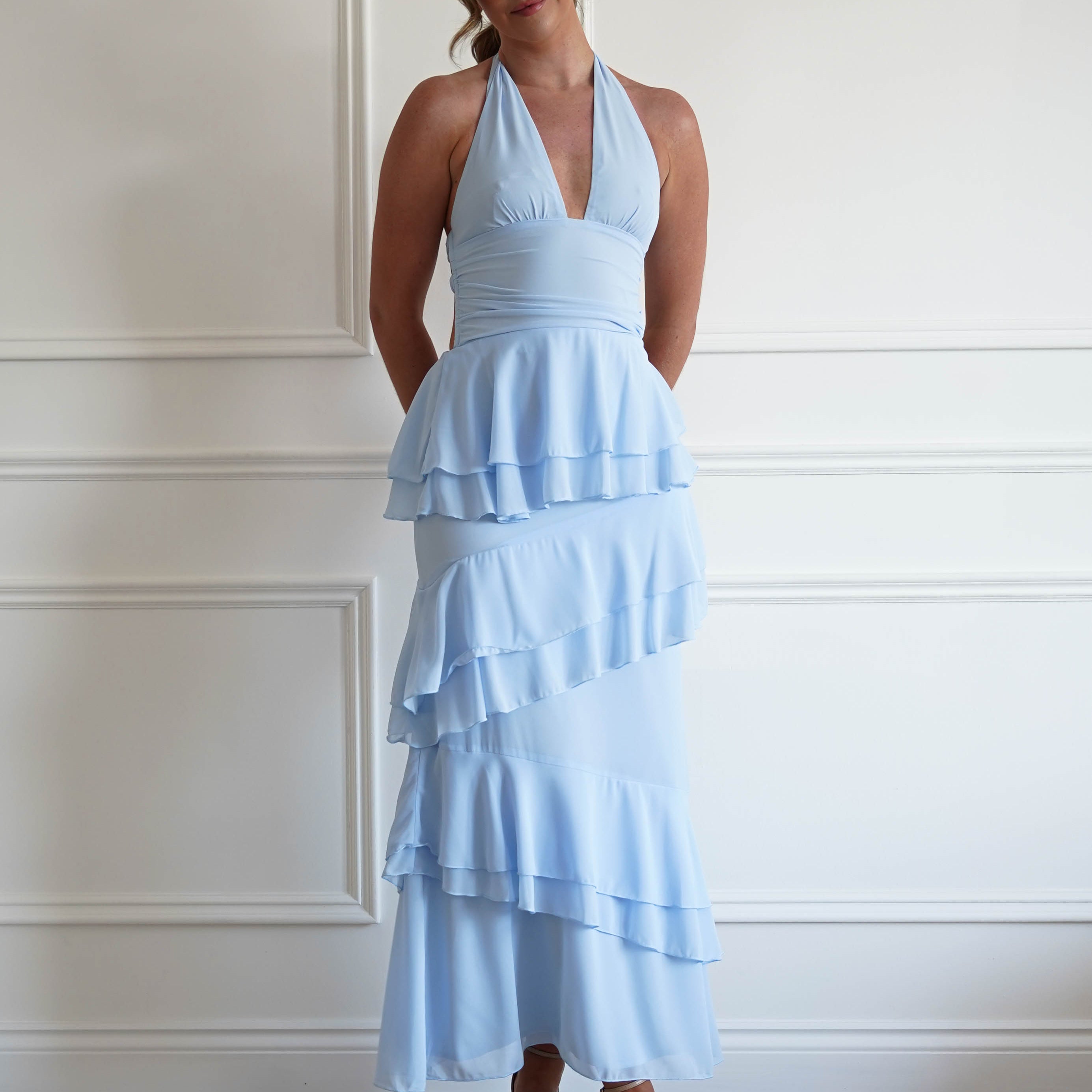 Woman wearing a light blue ruffled dress against a white paneled wall.