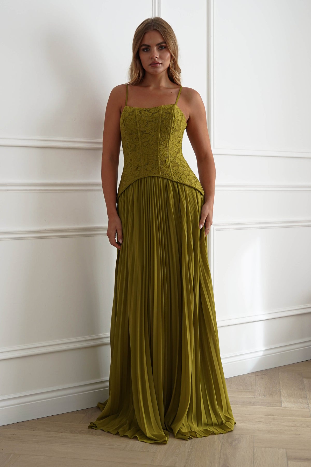 A woman modeling a floor-length olive green gown with a structured lace corset bodice and a flowing pleated skirt, standing against a white paneled wall on a light wood floor.
