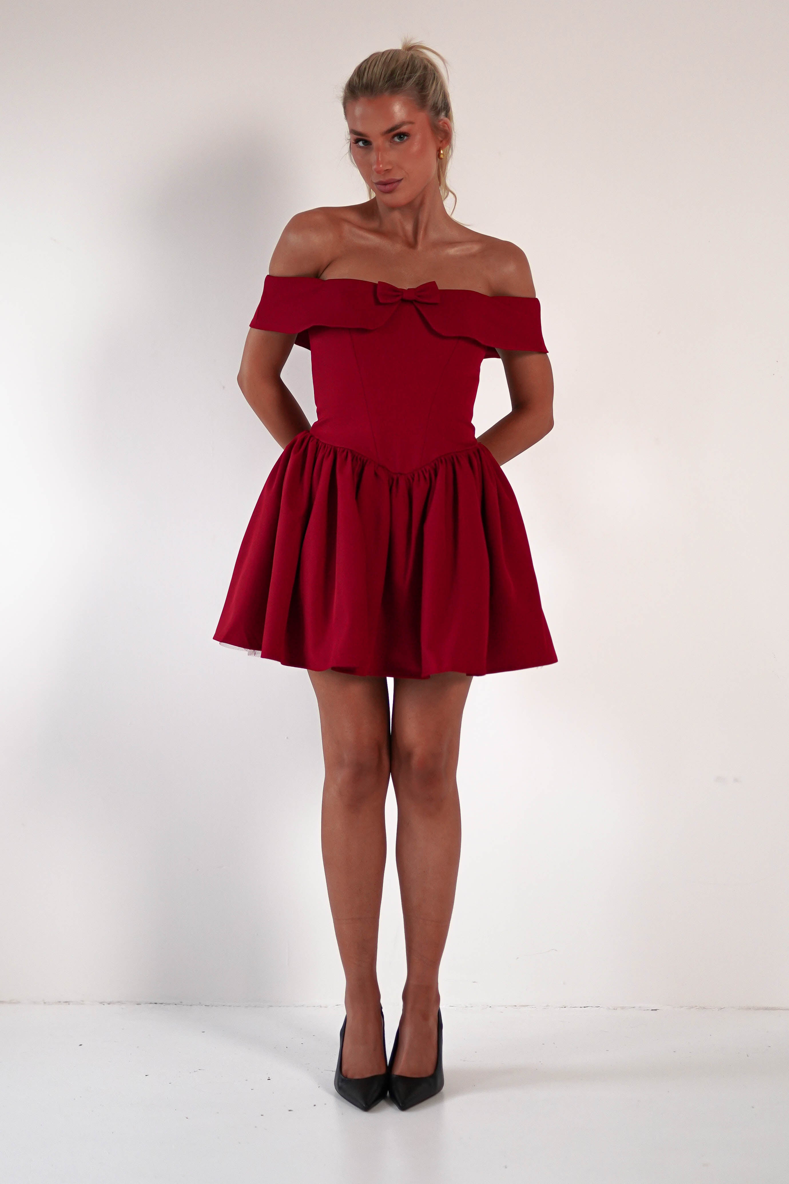 A woman in a red off-shoulder dress with a corset bodice stands against a plain background. The dress features a flared skirt, creating an elegant silhouette. She completes her look with black high heels and has her hair tied up gracefully.