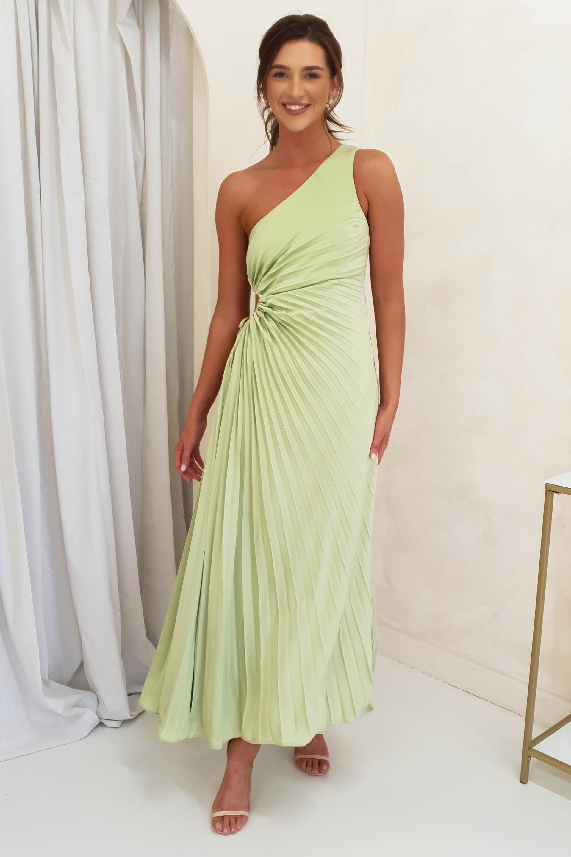 A woman wearing a light green one-shoulder pleated maxi dress stands smiling in a minimalist room with light walls and a white curtain—an elegant look perfect for wedding guests or formal events.