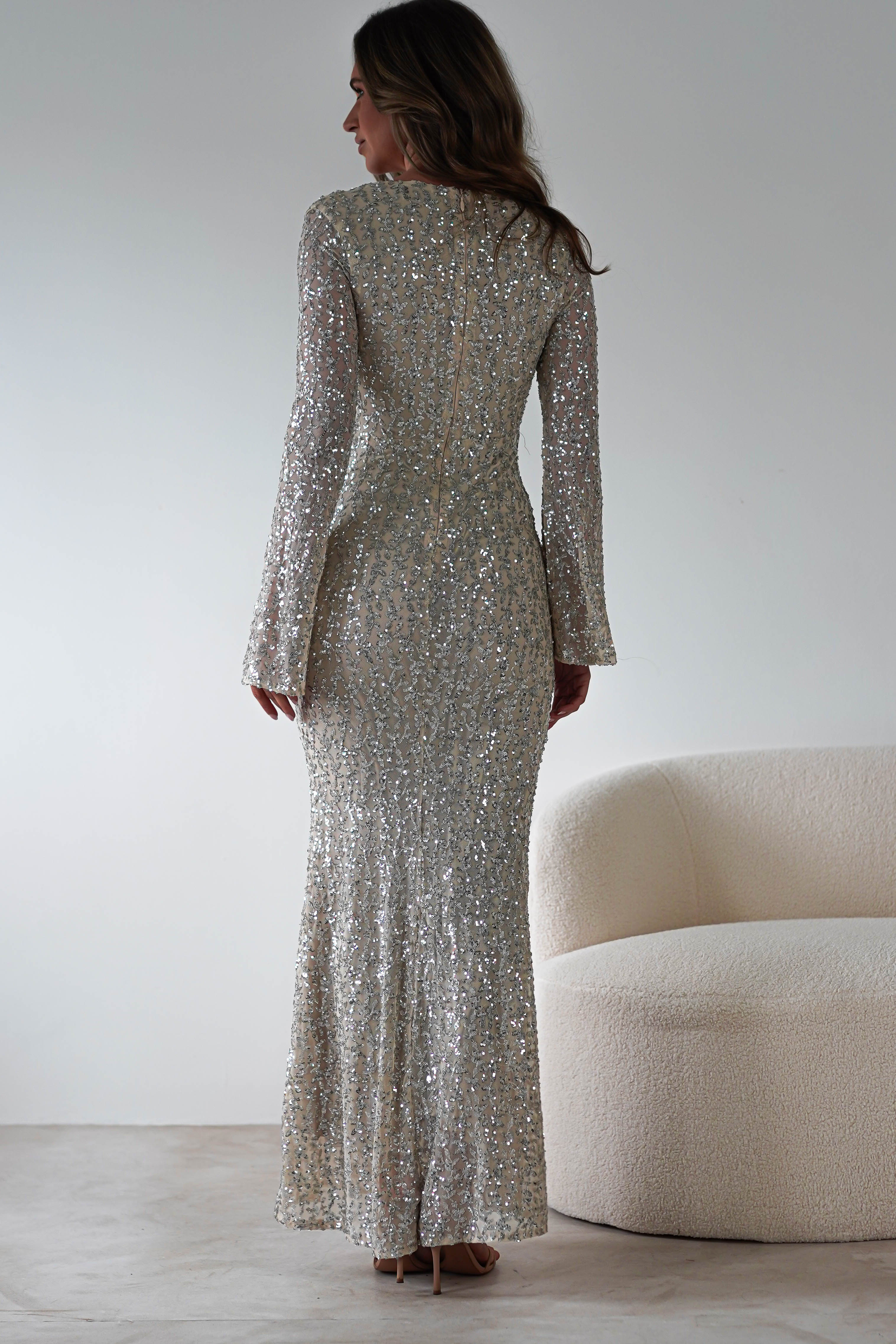 A woman in a sparkling, floor-length silver sequined gown stands facing away. The dress features long sleeves and a fitted silhouette, perfect for a wedding guest. She is next to a plush, light-colored chair against a minimalist white background.