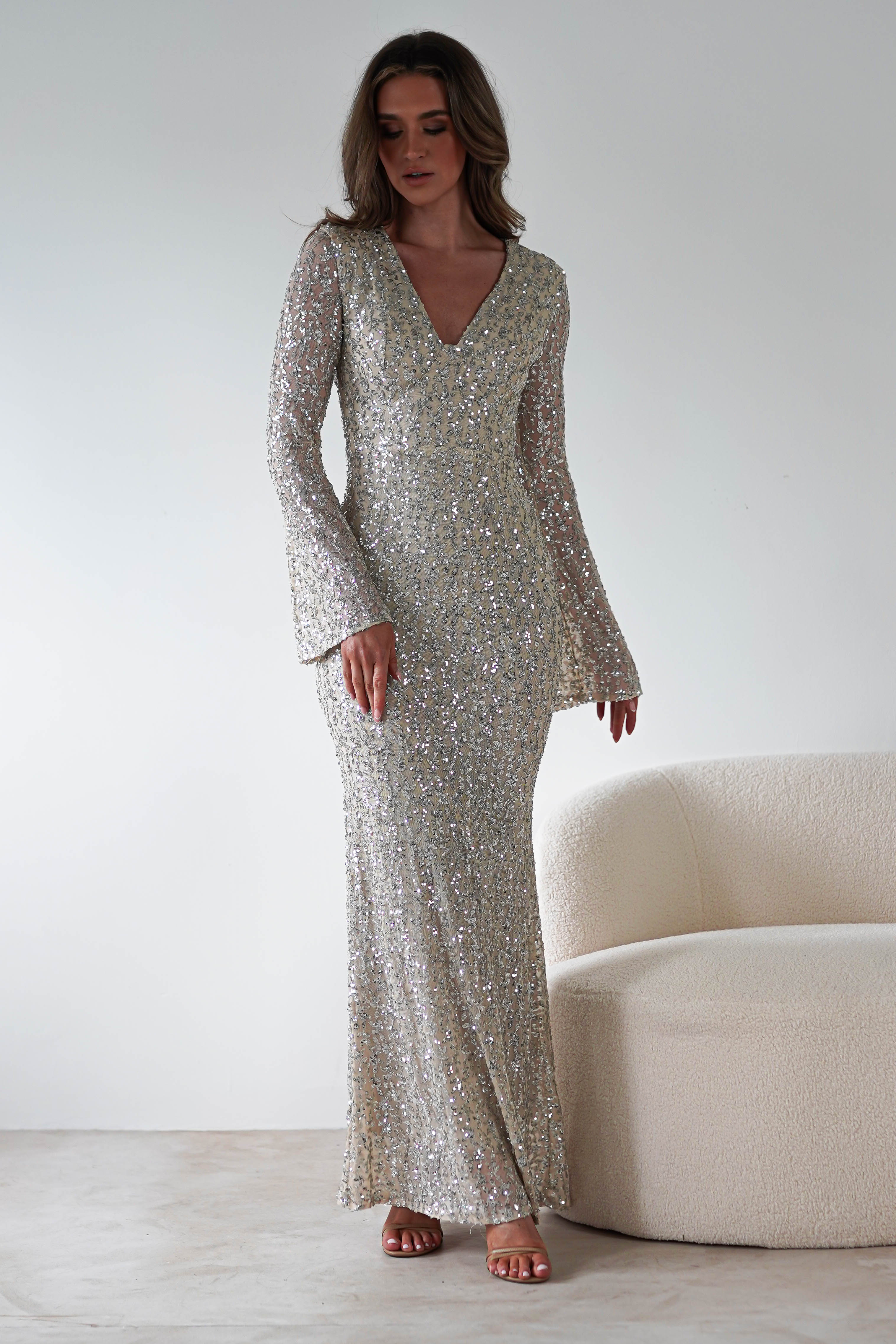 A person stands against a plain background, wearing a nude maxi party dress adorned with shimmering silver and sparkling sequins. The dress features long sleeves and a V-neckline. Their long hair flows elegantly as they pose beside a light-colored couch.