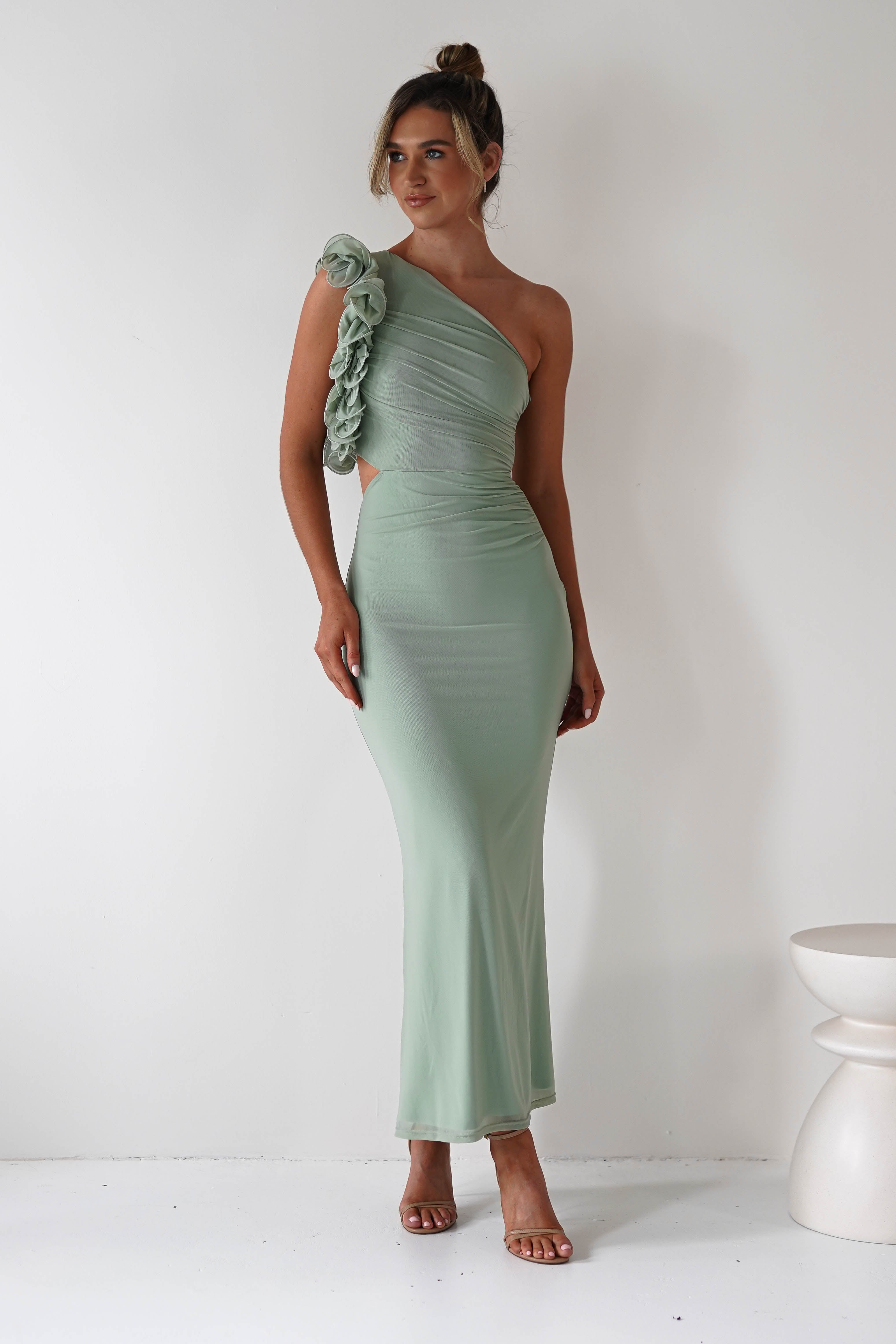 A woman in a mint green, one-shoulder midaxi dress with floral and ruffle features stands against a white background. Her hair is styled in a bun, she wears strappy heels, making for the perfect wedding guest outfit. A white stool is visible to the side.