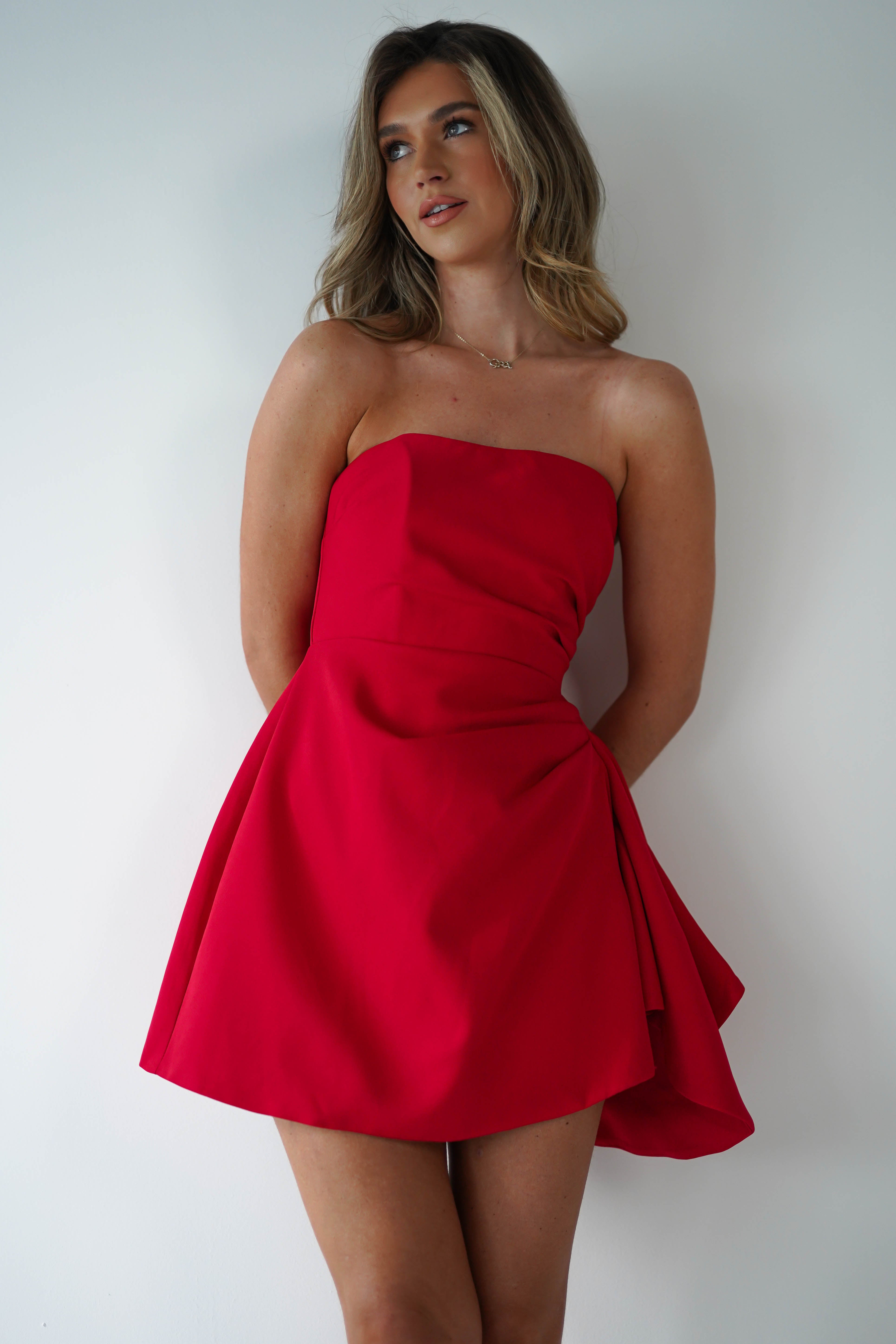 A woman in a strapless mini dress, perfect for a wedding guest, stands against a white background, looking thoughtfully to the side. Her hands are behind her back, and her long hair cascades over her shoulders.