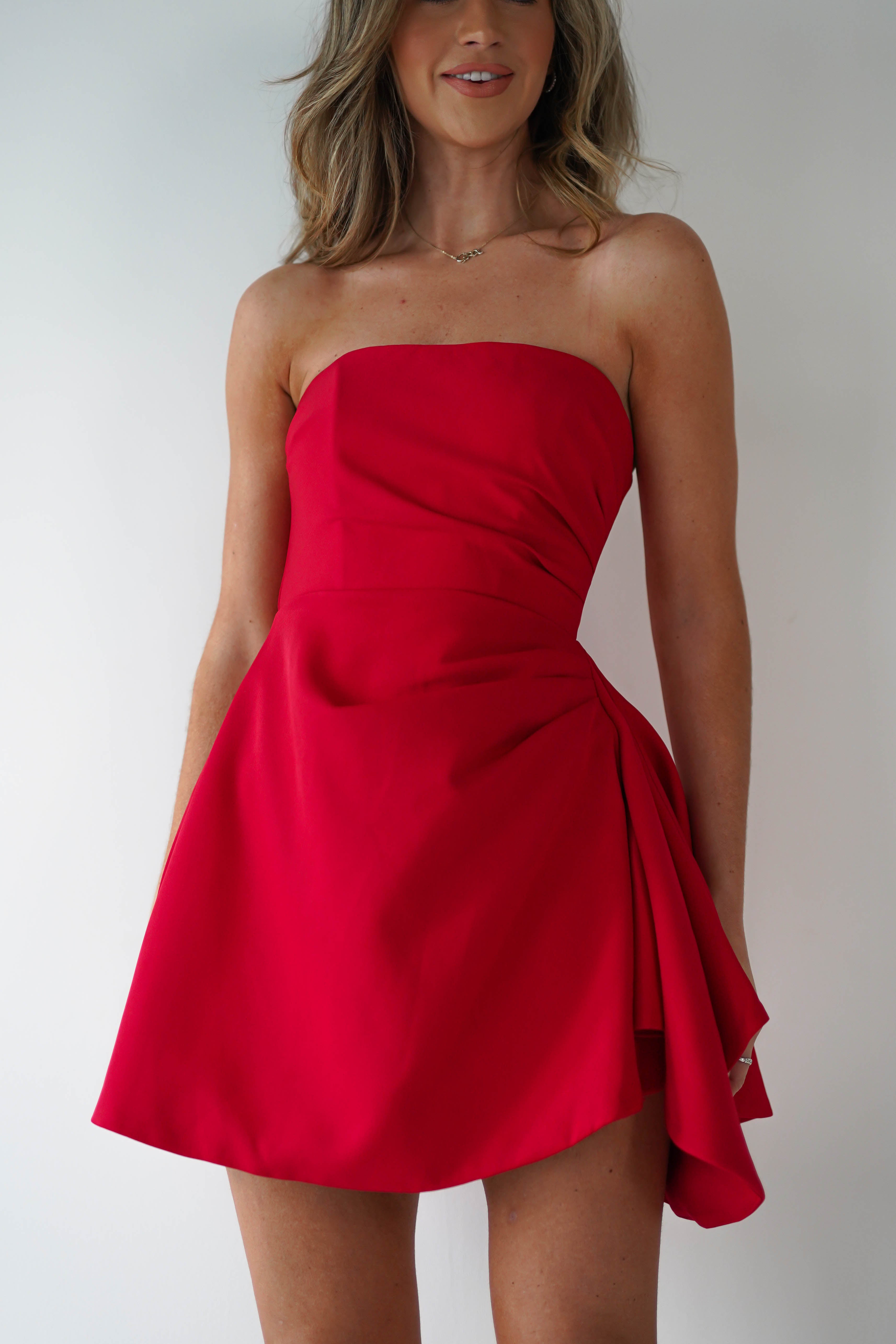 A woman stands against a plain white background, her long hair cascading down. Shes elegantly dressed in a strapless mini dress that flares slightly at the waist, perfect for any party. The red dress features subtle draping on the side, adding a touch of sophistication to her ensemble.