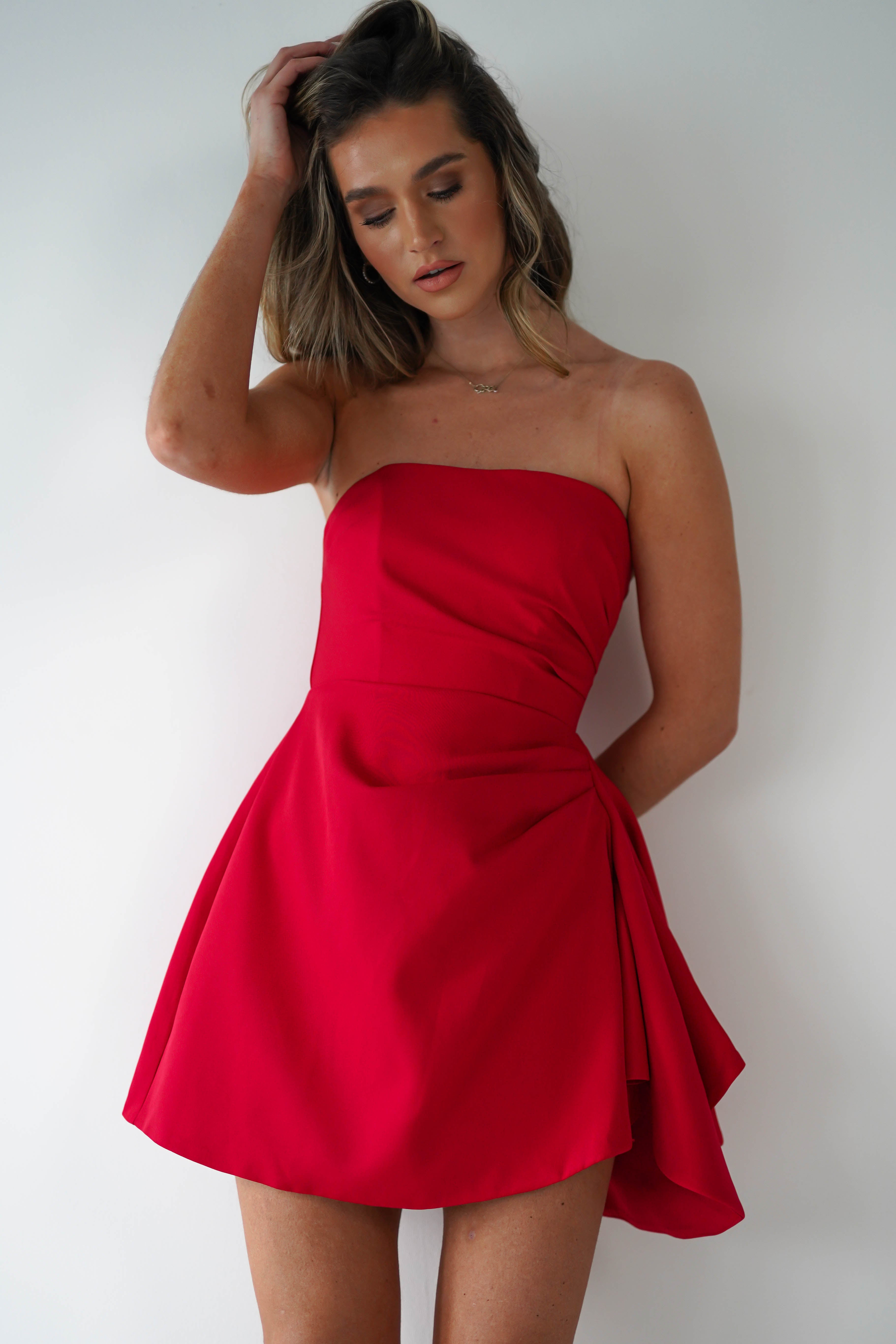 A woman stands against a white background, wearing a strapless red party dress with a flared skirt. She has shoulder-length hair and is touching her head with one hand while looking downwards.