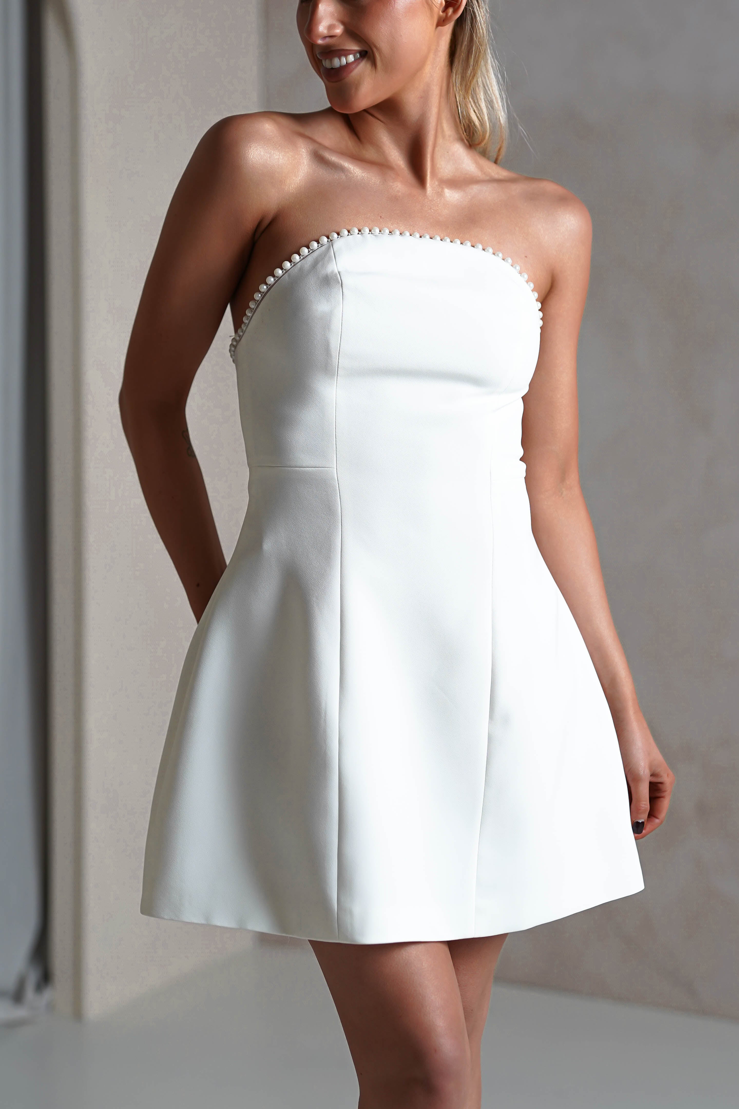 A woman wearing a strapless white party dress with pearl-like beads along the neckline. The fitted bodice and flared skirt enhance her look as she stands with her arm behind her, smiling and posing indoors against a soft background.