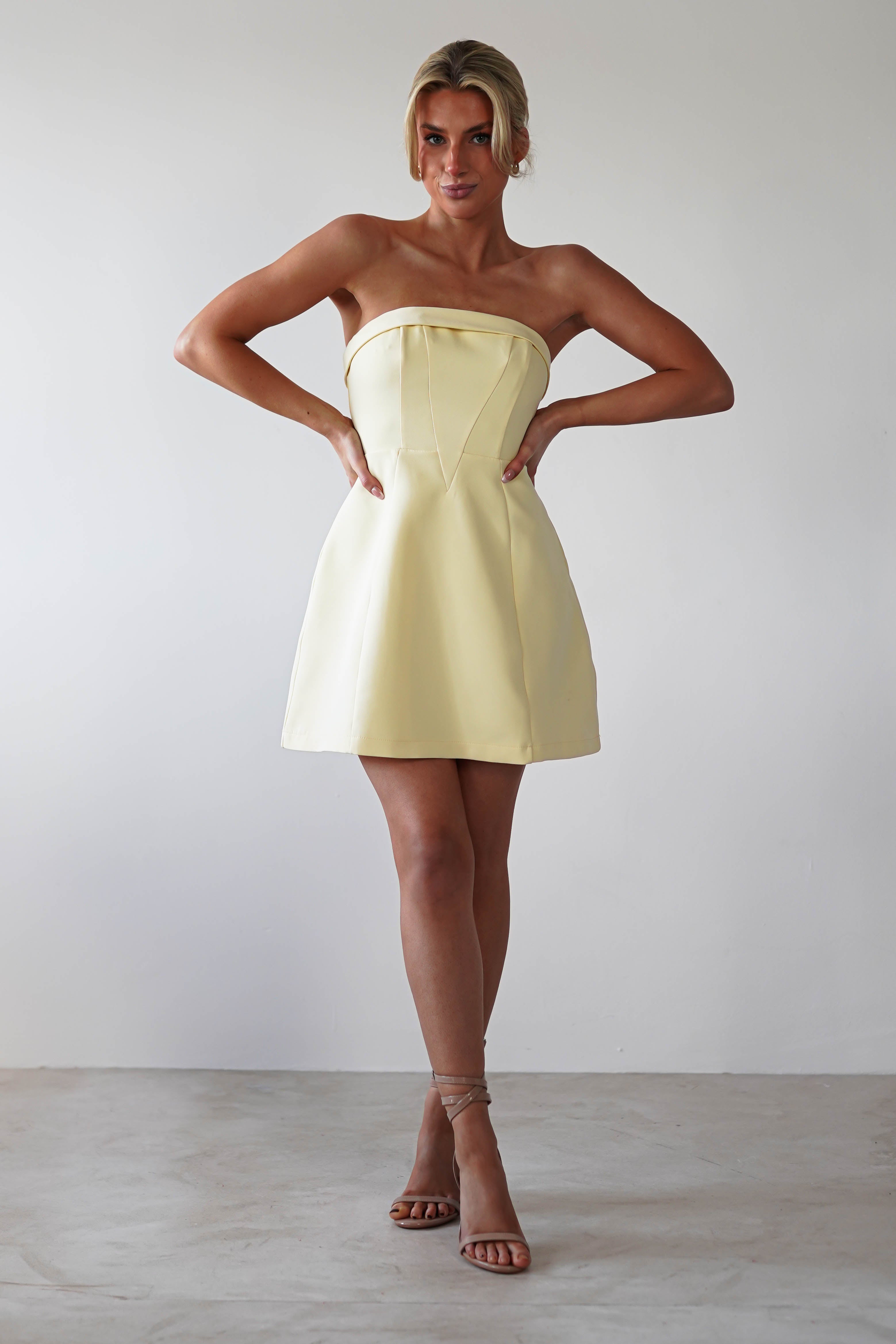 A person stands confidently wearing a strapless, light yellow party dress. Their hands rest on their hips as they showcase strappy, neutral-colored heels. The plain, light-colored background enhances the elegance of this wedding guest dress.