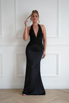Woman wearing a black bodycon halterneck maxi dress with a plunging V-neckline, ruched waist detailing, and a fitted floor-length silhouette, perfect for formal events, black-tie occasions, and evening wear.