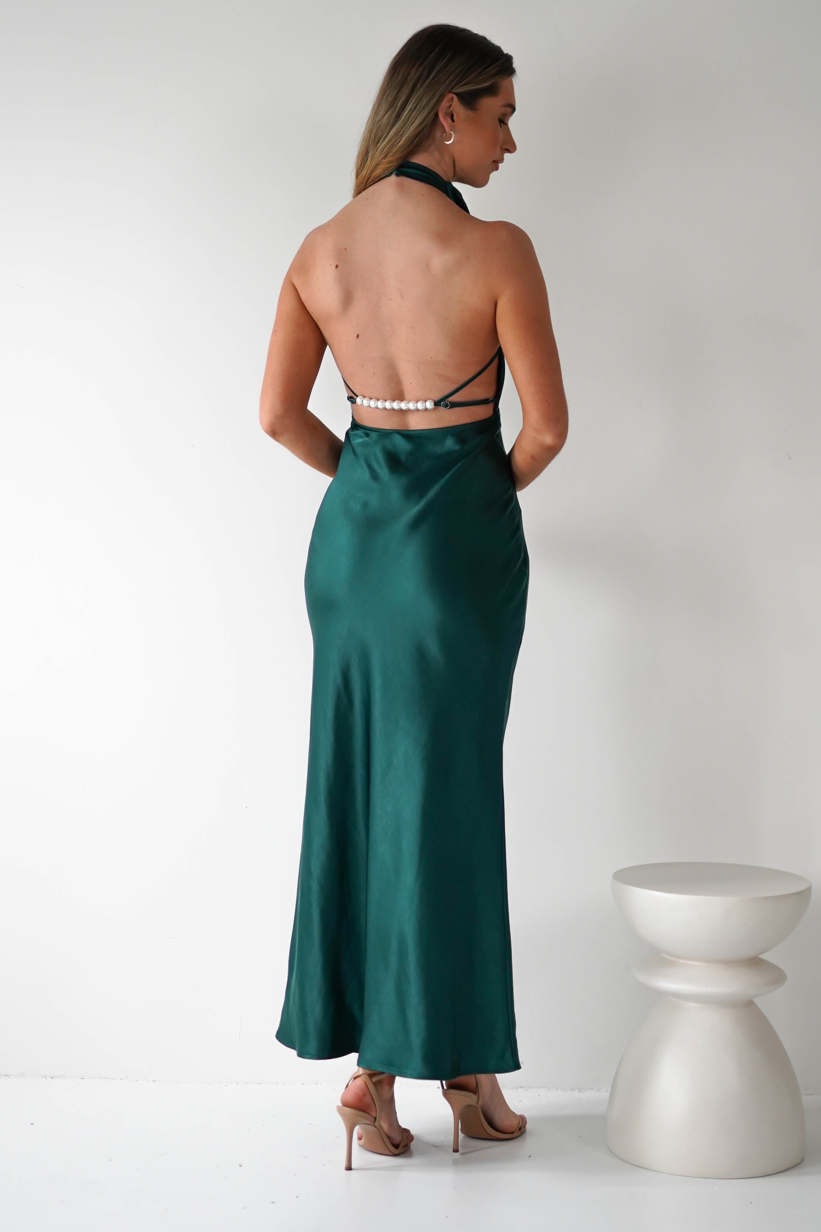 A woman in an emerald green satin gown stands facing a white wall, showcasing its open-back design with elegant pearl detail. Her beige high heels complement the ensemble, as a white, modern sculptural stool sits nearby.