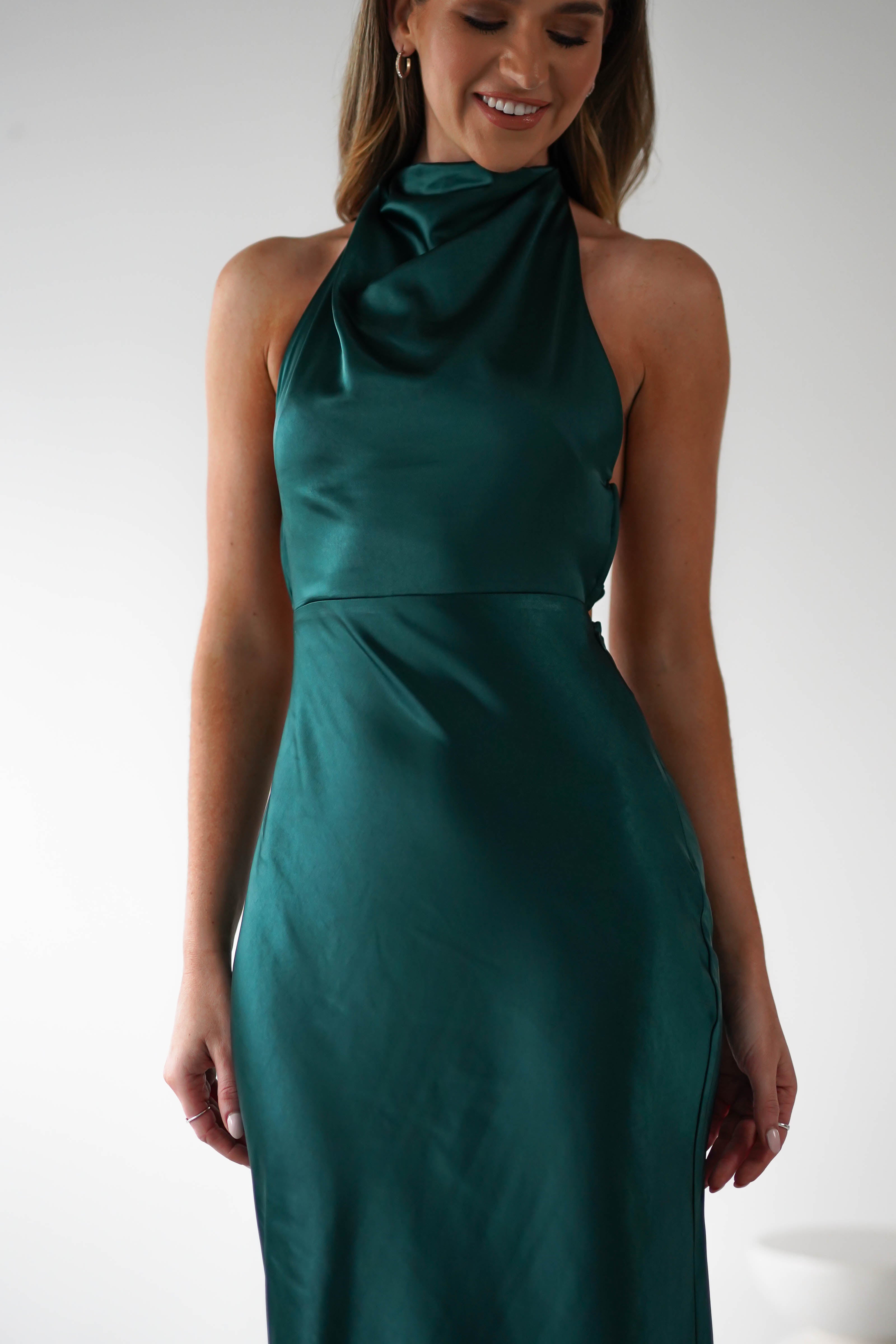 A woman wearing a sleek, sleeveless emerald green satin dress with an open-back design smiles. The dress features a high neck and an elegant, fitted silhouette. She is posing against a plain white background.