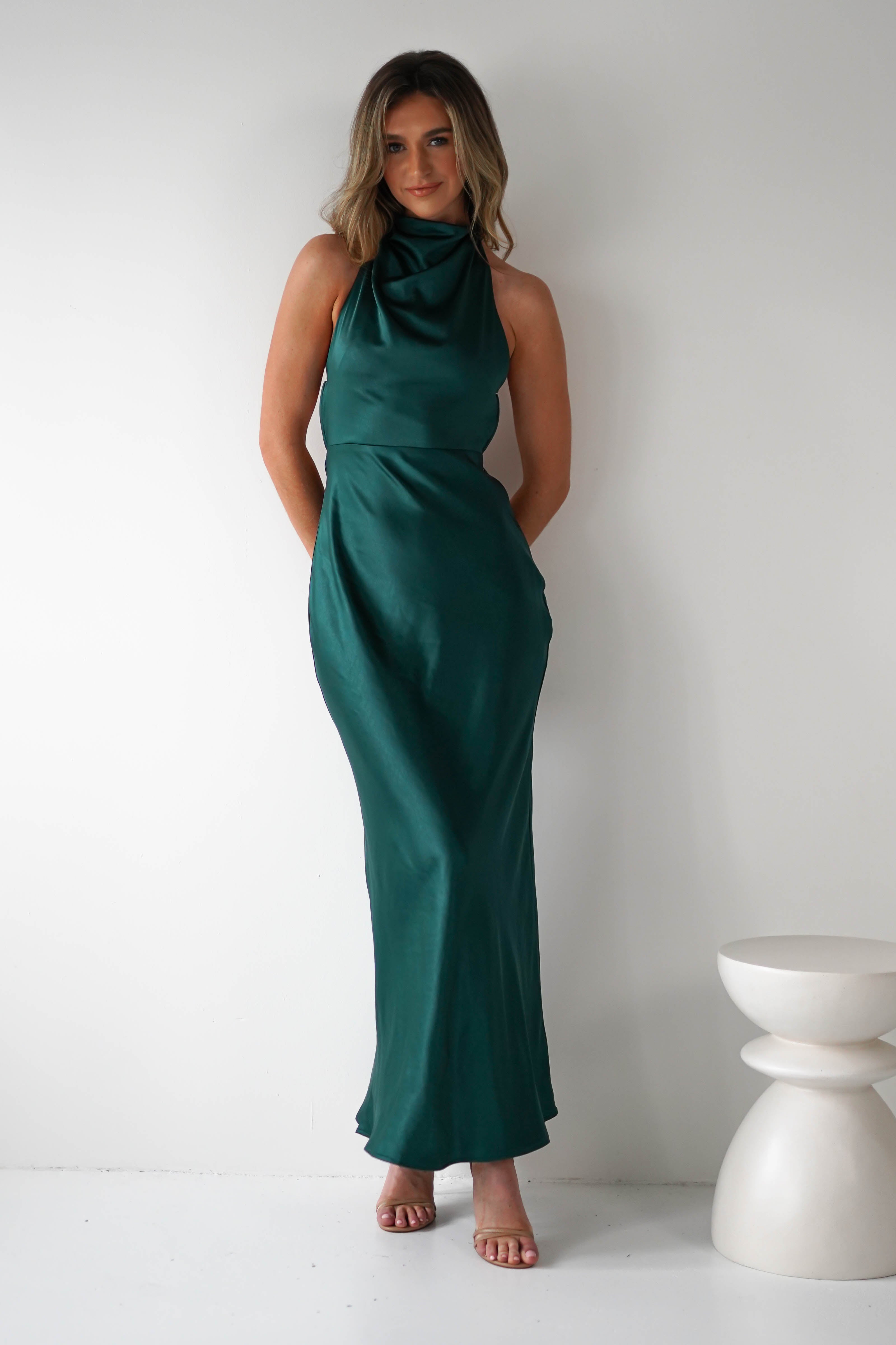 A woman stands against a white wall in a sleeveless, dark green satin dress featuring an elegant open-back design. Her hands rest casually behind her back as she gazes at the camera. Beside her is a small, white modern stool.