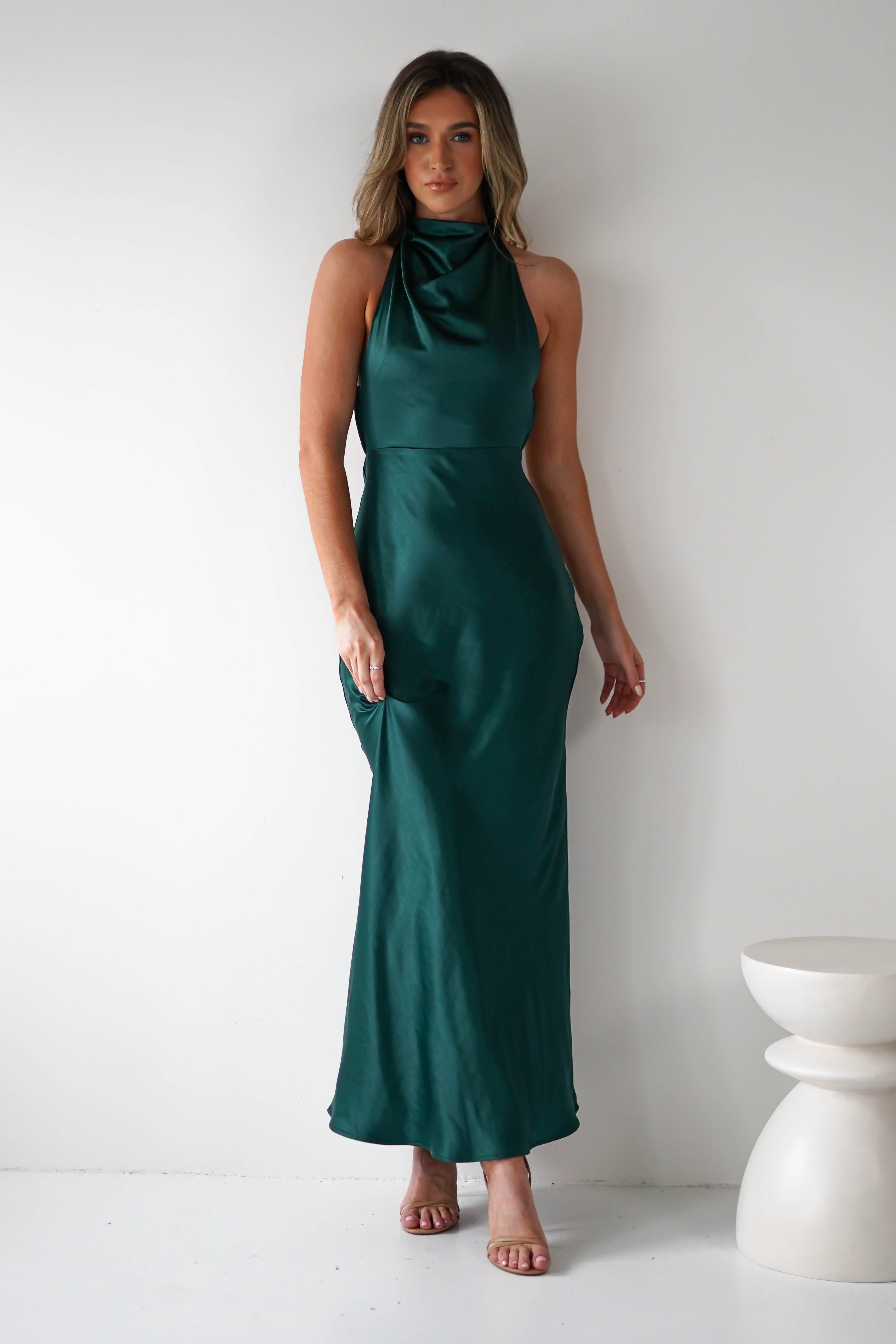 A woman stands gracefully in an open-back, green sleeveless gown. The dress is long and form-fitting with exquisite pearl detail. Her long, wavy hair cascades down as she completes the look with heels against a stark white wall and a small pedestal to her right.