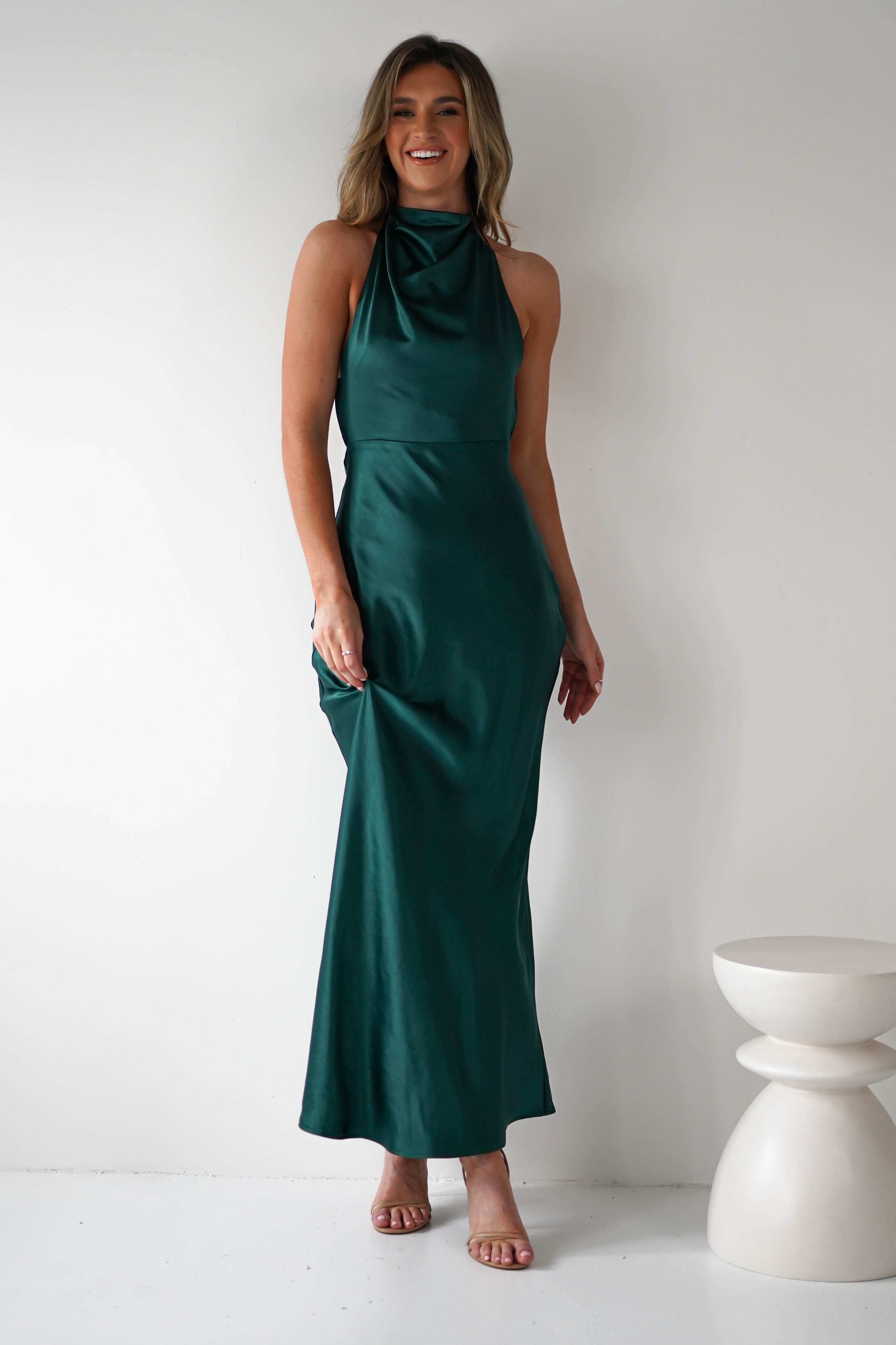 A woman in a long, dark green satin dress with pearl detail stands against a white background, smiling. The dress features a high neckline and flowing silhouette. She holds the dress with one hand and wears nude high-heeled sandals. A round stool is nearby.