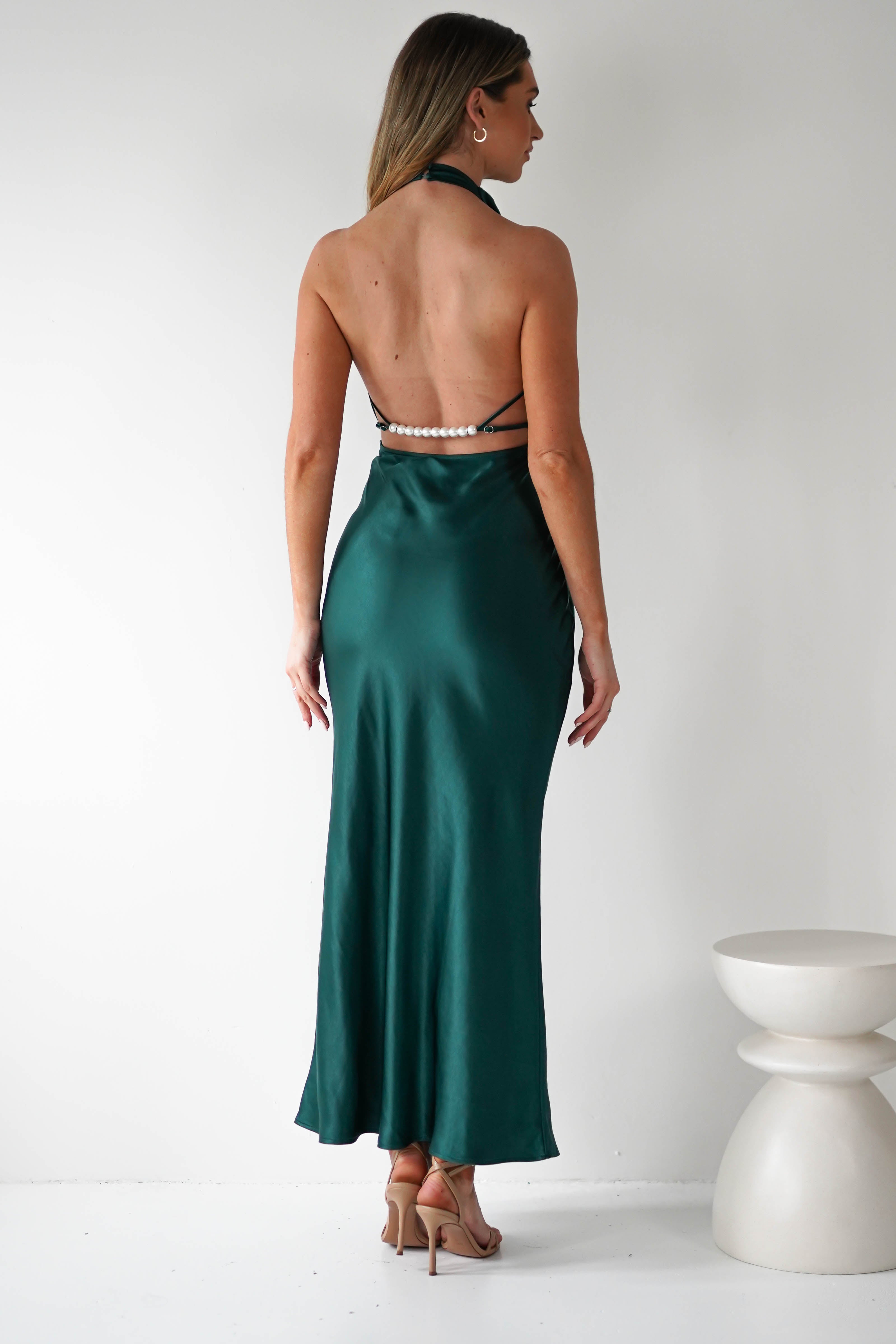 A woman stands facing away, showcasing the open-back design of her dark green satin dress with pearl details on the straps. She is barefoot on a white floor beside a rounded side table against a plain white wall.