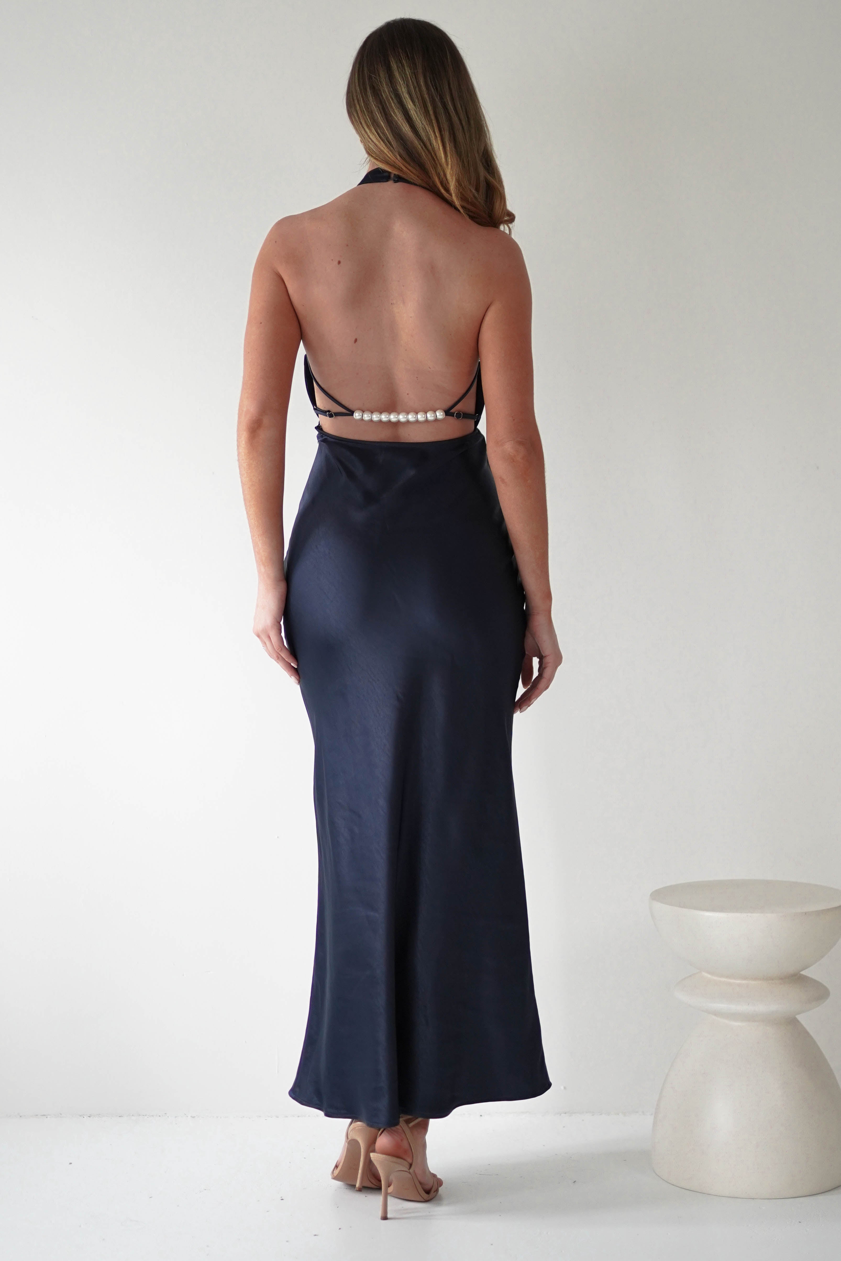 A woman with long hair faces away in a sleek black satin dress featuring an open-back design and pearl details. The floor-length gown is paired elegantly with nude high heels. She stands in a minimalist setting, complemented by a light-colored abstract pedestal nearby.