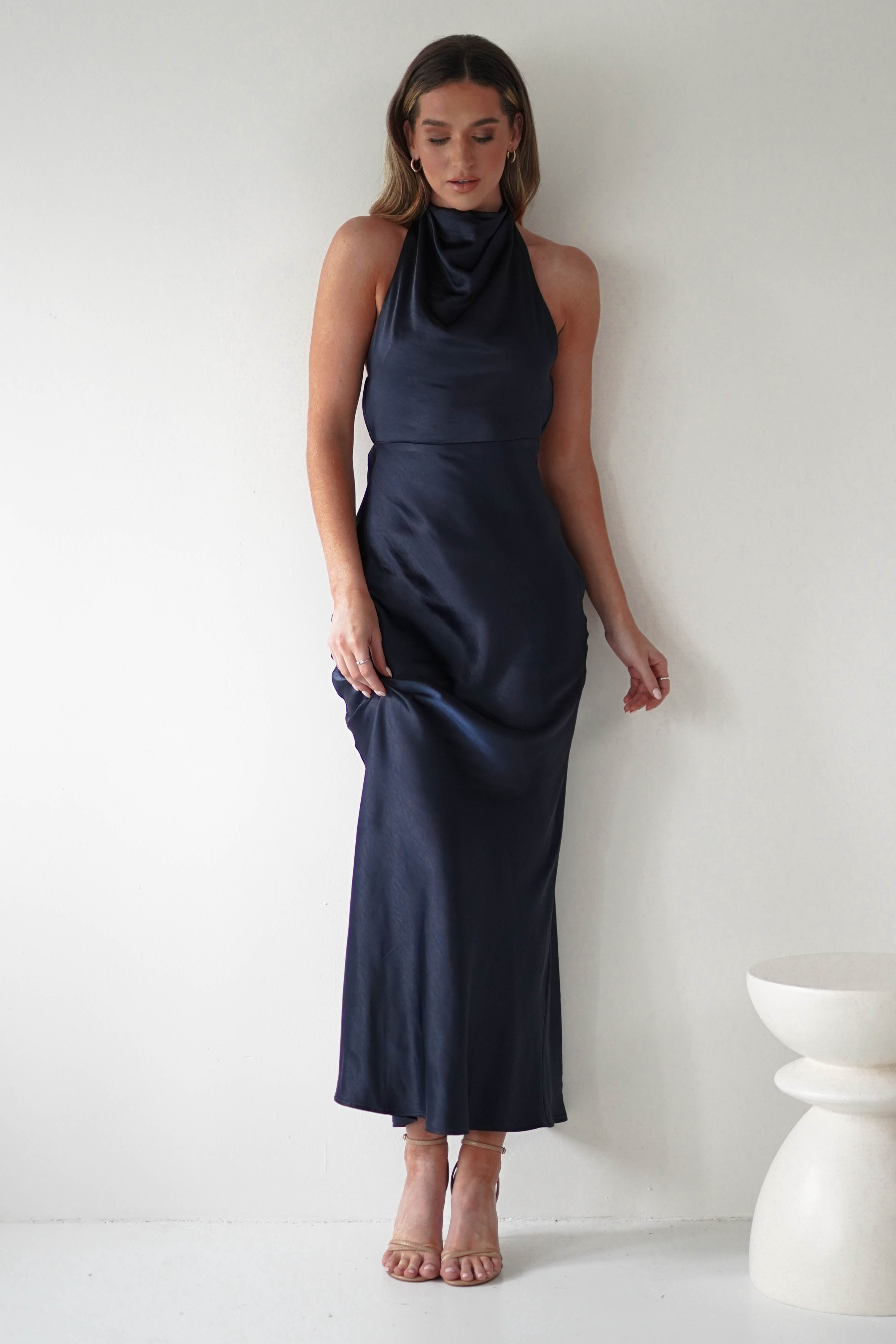 A woman stands against a white wall, wearing a sleek, dark blue satin dress with an open-back design and halter neckline. She has light brown hair and is looking down. Her right hand lightly grasps the fabric of the dress. A white, round stool is beside her.