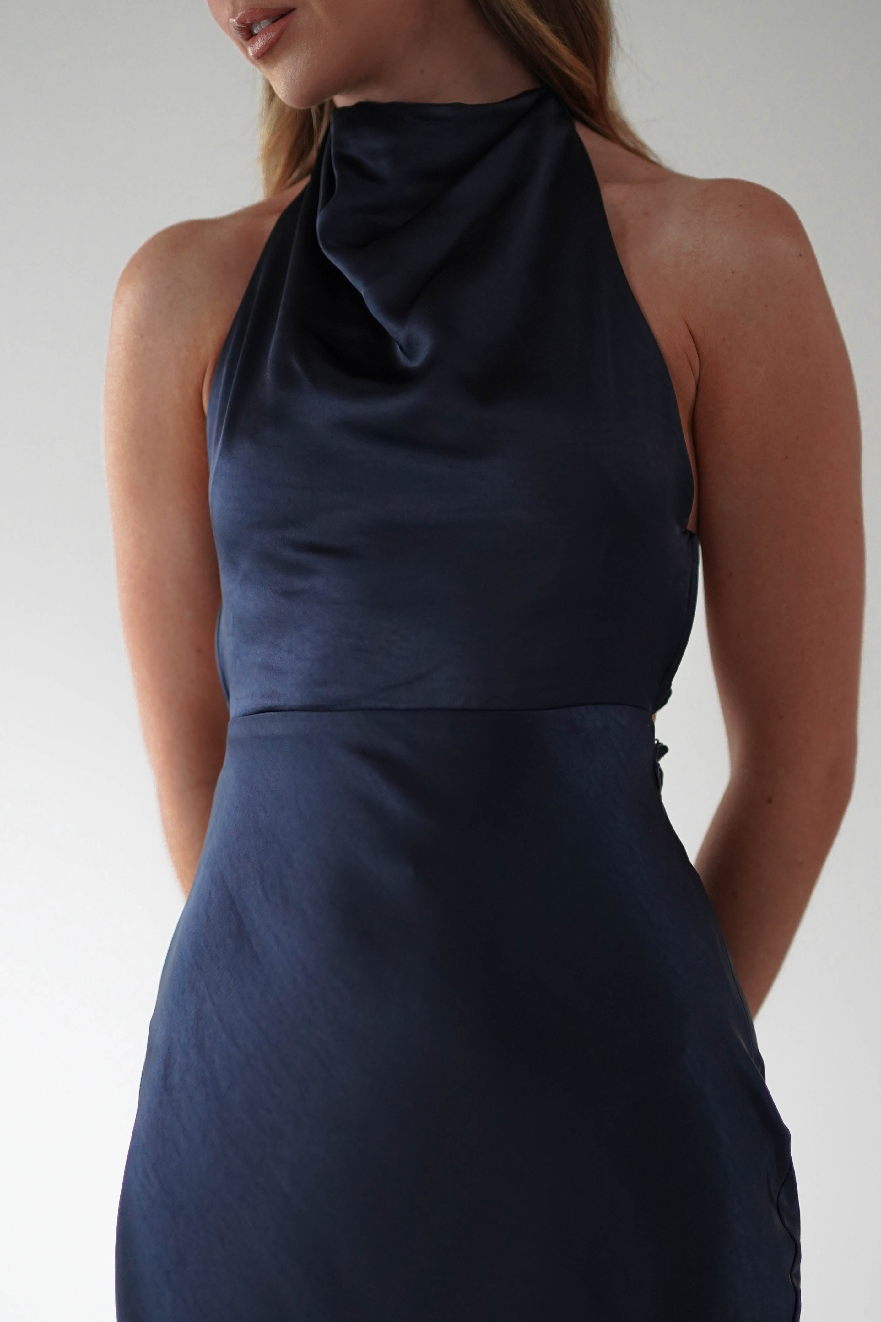 A person wearing a sleeveless, black satin dress with a halter neck and an elegant open-back design stands against a plain background. Their head is partially turned, showing long hair and a hint of a smile. The focus is on the upper half of the dress.