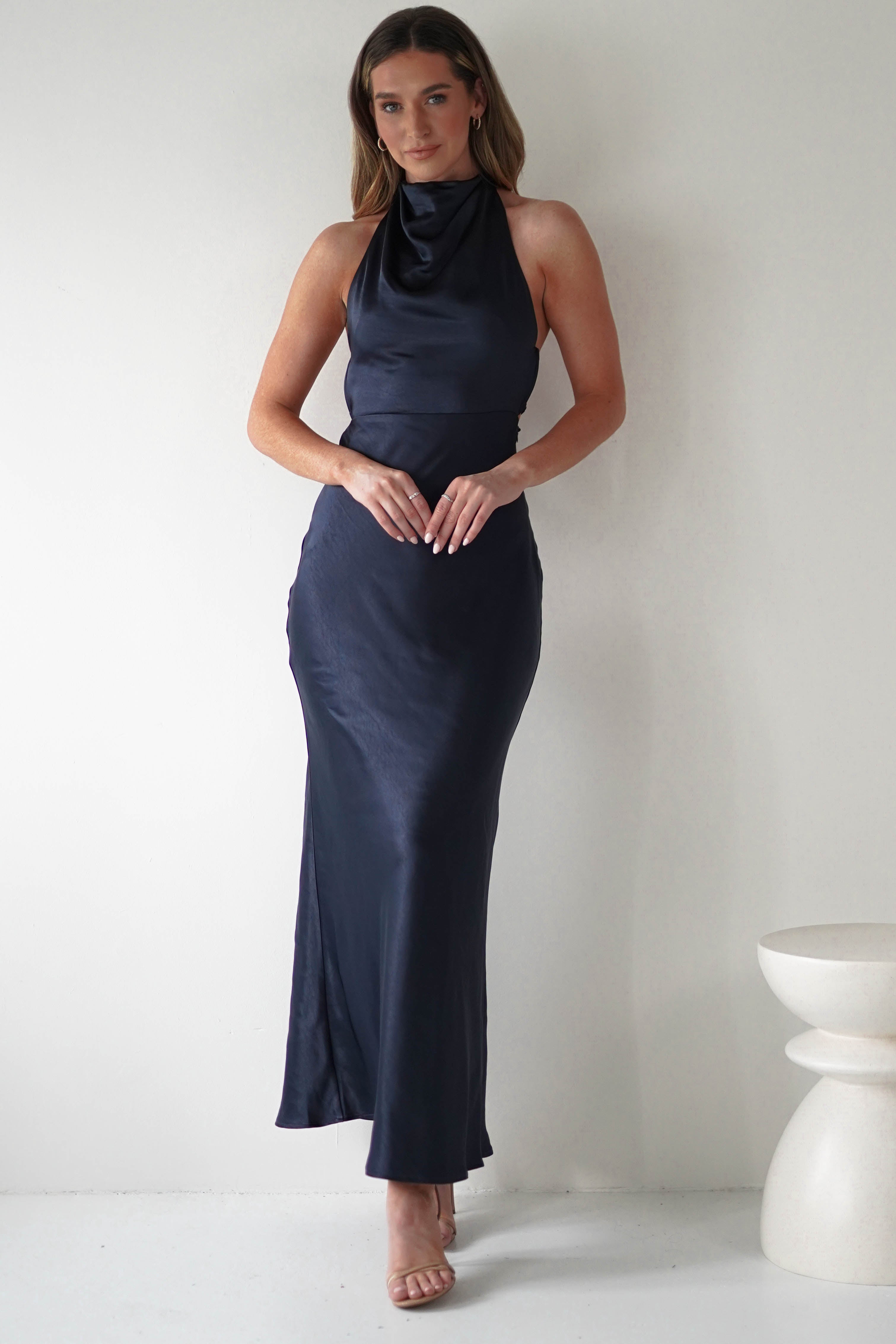 A person stands against a plain wall, wearing a sleek, dark blue halter-neck gown with an elegant open-back design. Their hands are gently clasped in front. They have long hair and are wearing open-toed heels. A small white stool is partially visible on the right.