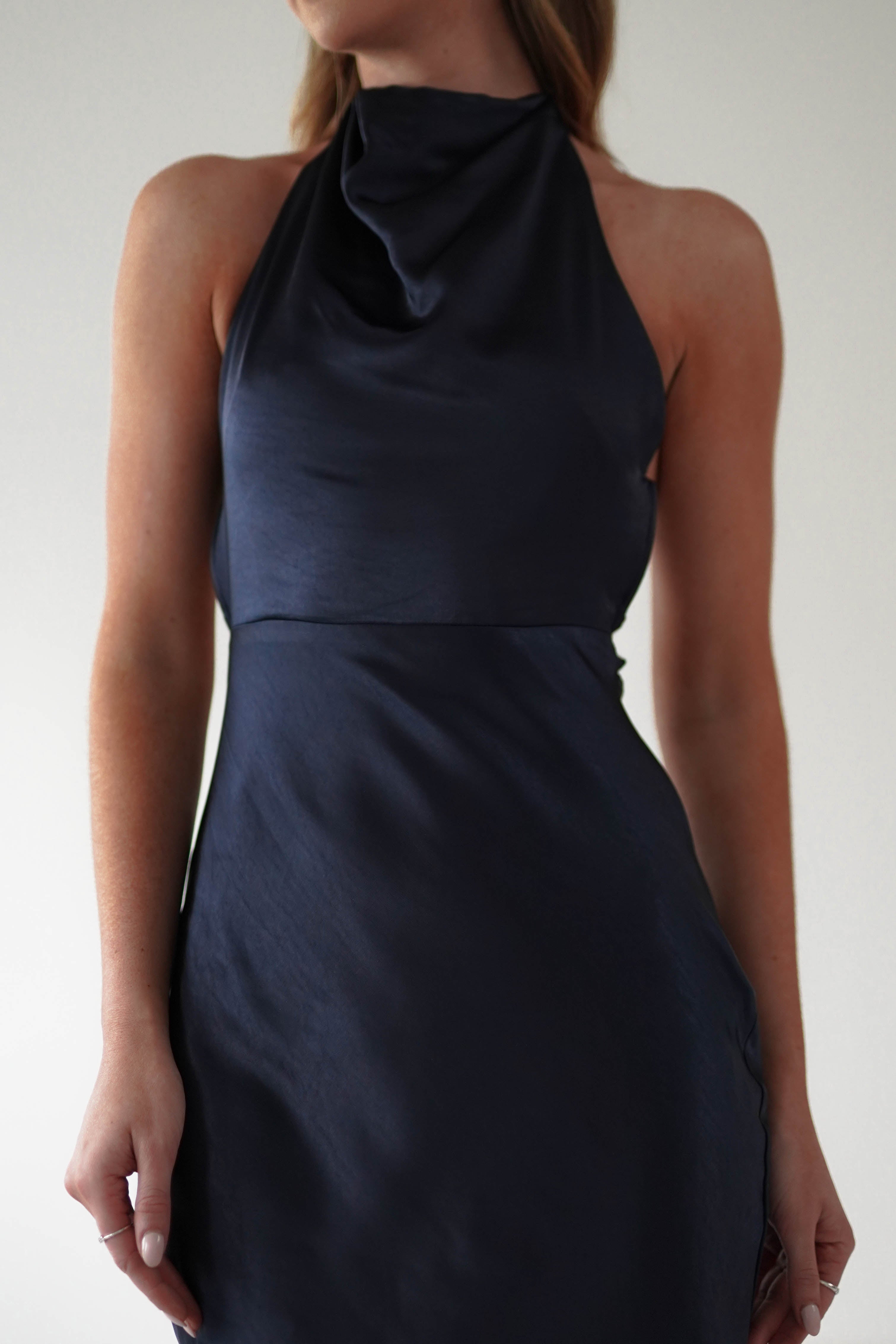 A woman wearing a sleeveless black satin dress with an open-back design stands against a neutral background, captured from shoulders to mid-thigh. Her arms are relaxed by her sides, and her hair falls straight over her shoulders.