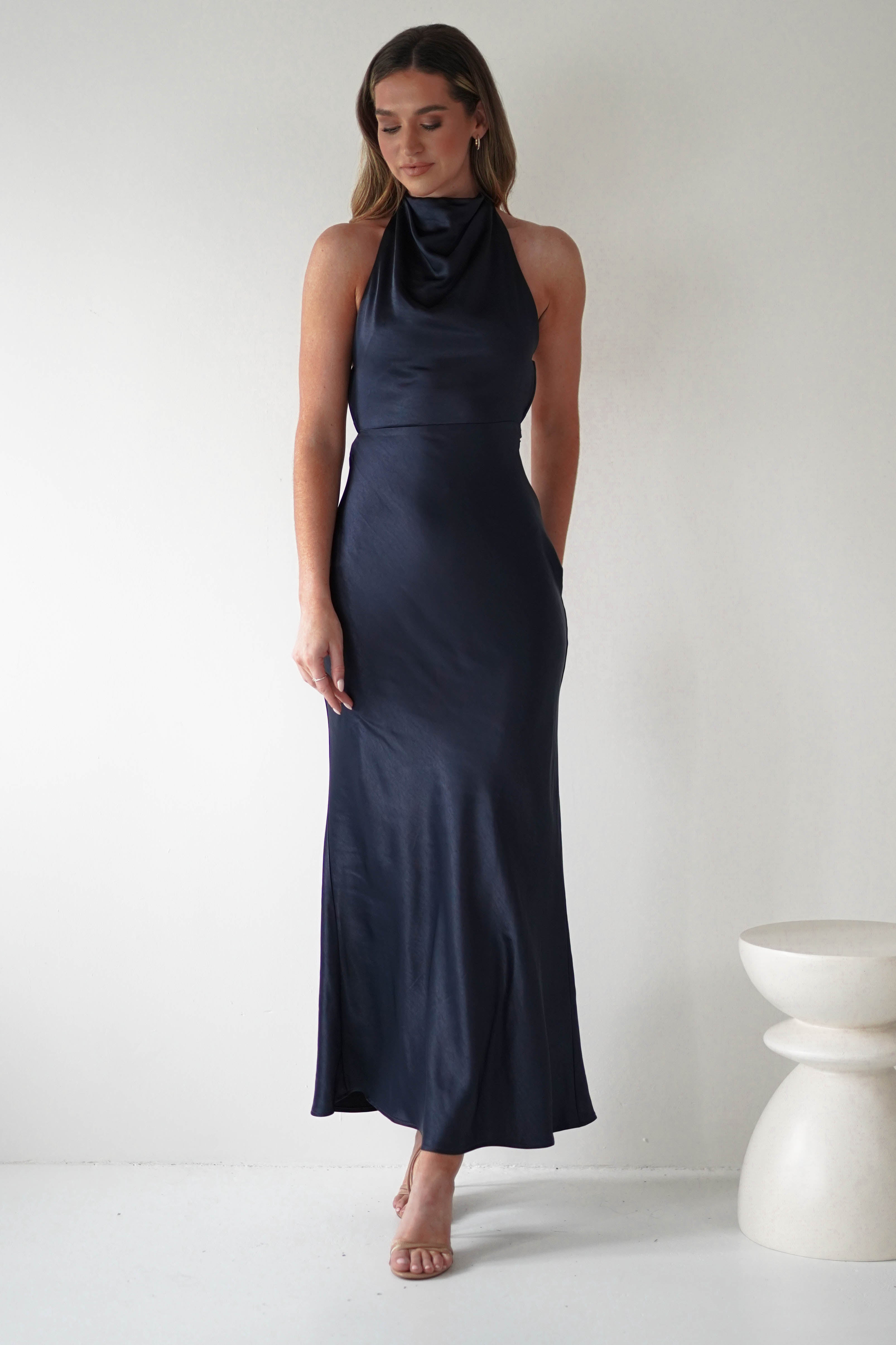 A woman stands in front of a plain white background, wearing a sleek, dark blue halter-neck evening gown with pearl detail. She has long hair and is wearing minimal accessories. A small round table is partially visible beside her.