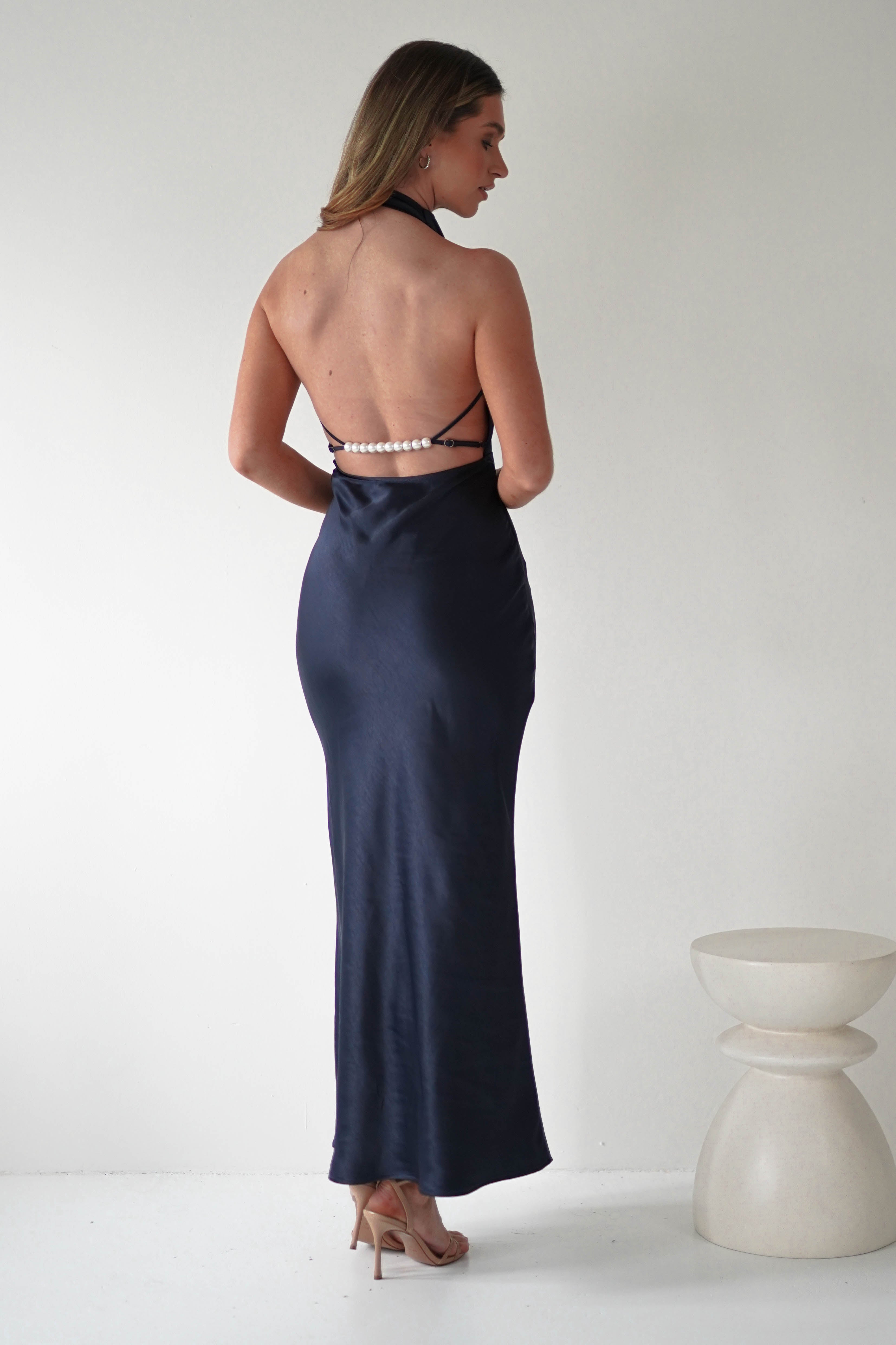 A woman stands in a navy blue backless gown with pearl accents and spaghetti straps. She is facing away, showcasing the open-back design and wearing beige high heels. The background is a minimalist white setting with a small, smooth, white sculpture on the right.