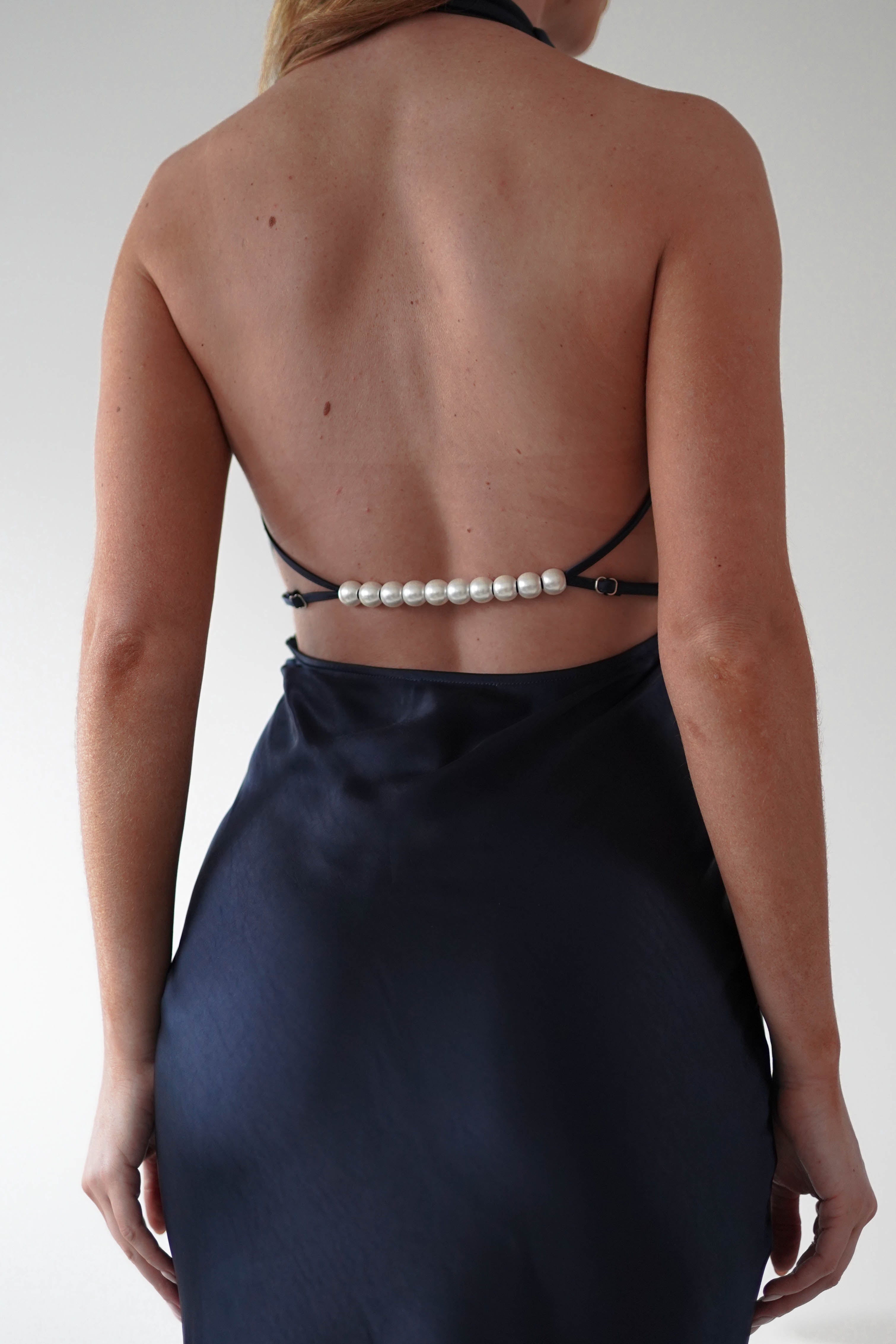 The image shows a person standing with their back to the camera, wearing a stunning black satin dress. The open-back design is elegantly enhanced by a string of pearl details cascading across the lower back.