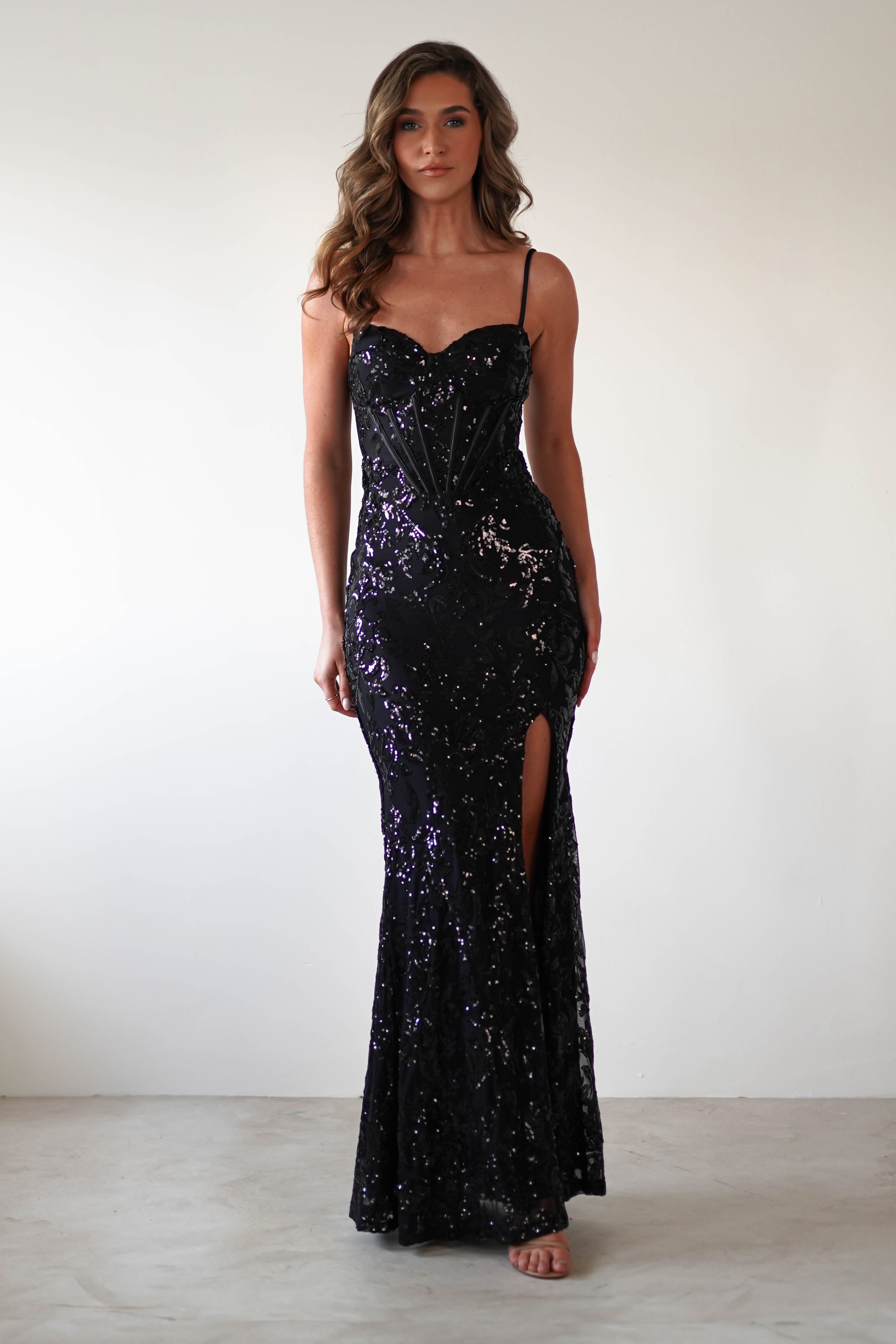 A woman wearing a black sequined gown with spaghetti straps poses against a plain white background. The dress, adorned with exquisite sequin embroidery, features a sweetheart neckline and a thigh-high slit on one side. Her hair is styled in loose waves.