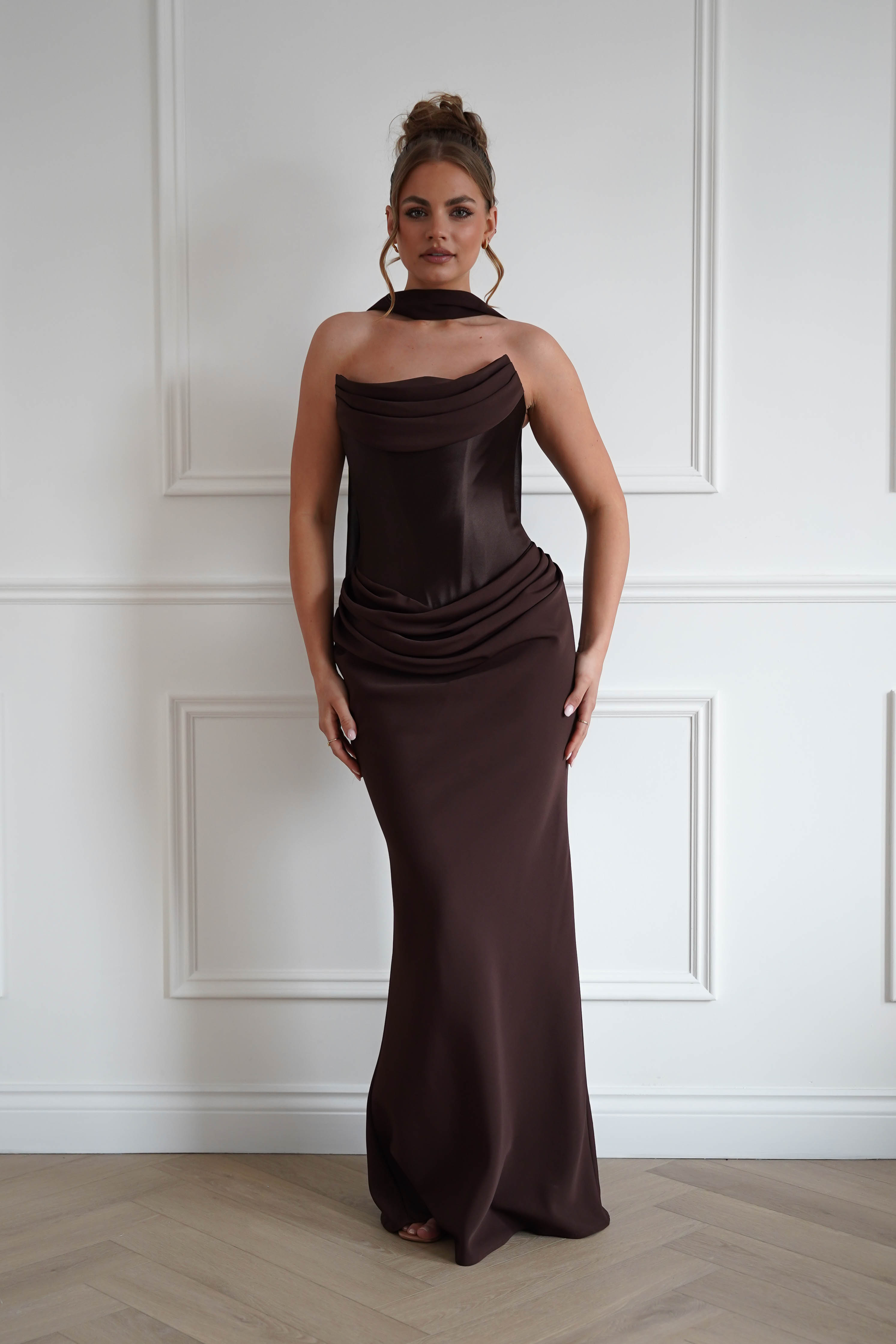 Woman wearing a chocolate brown strapless maxi dress with a draped neckline, ruched hip detailing, matching neck scarf, and fitted floor-length silhouette, ideal for weddings, black-tie events, galas, and formal evening occasions.