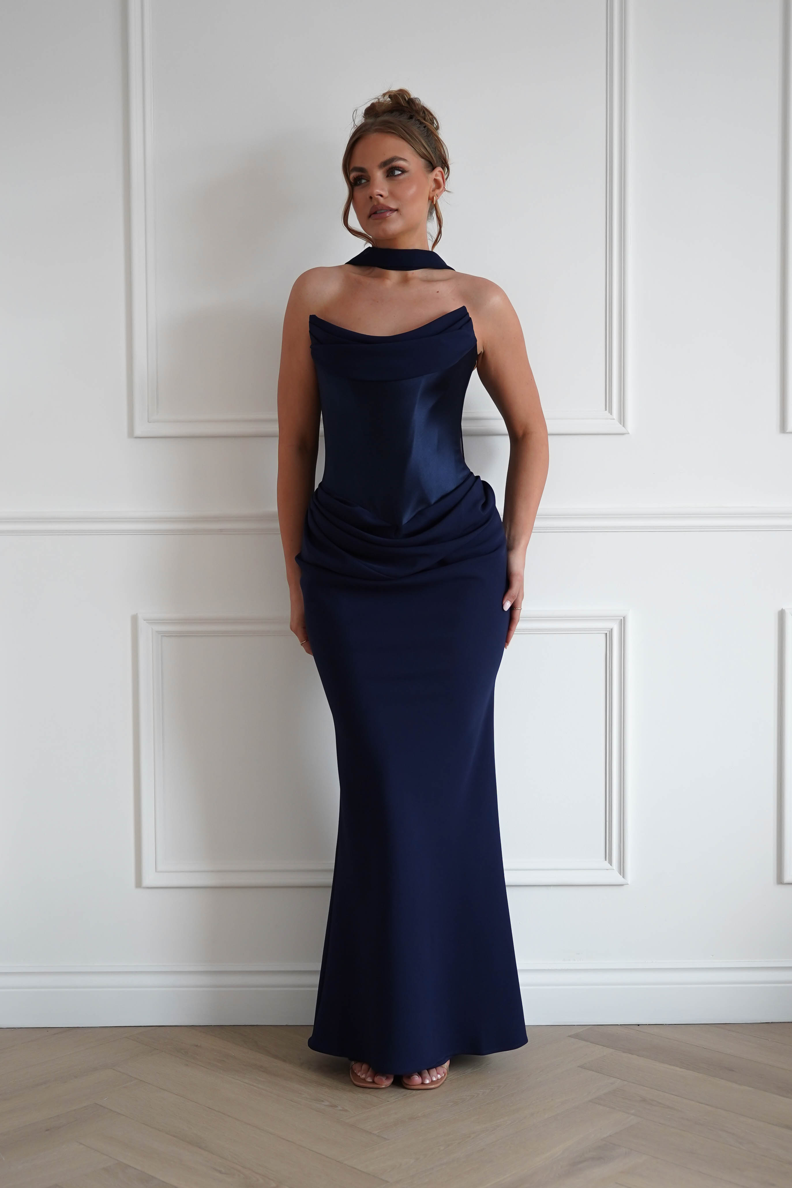 Woman wearing a navy strapless maxi dress with a draped neckline, ruched hip detailing, matching neck scarf, and fitted floor-length silhouette, perfect for black-tie events, weddings, galas, and formal evening occasions.