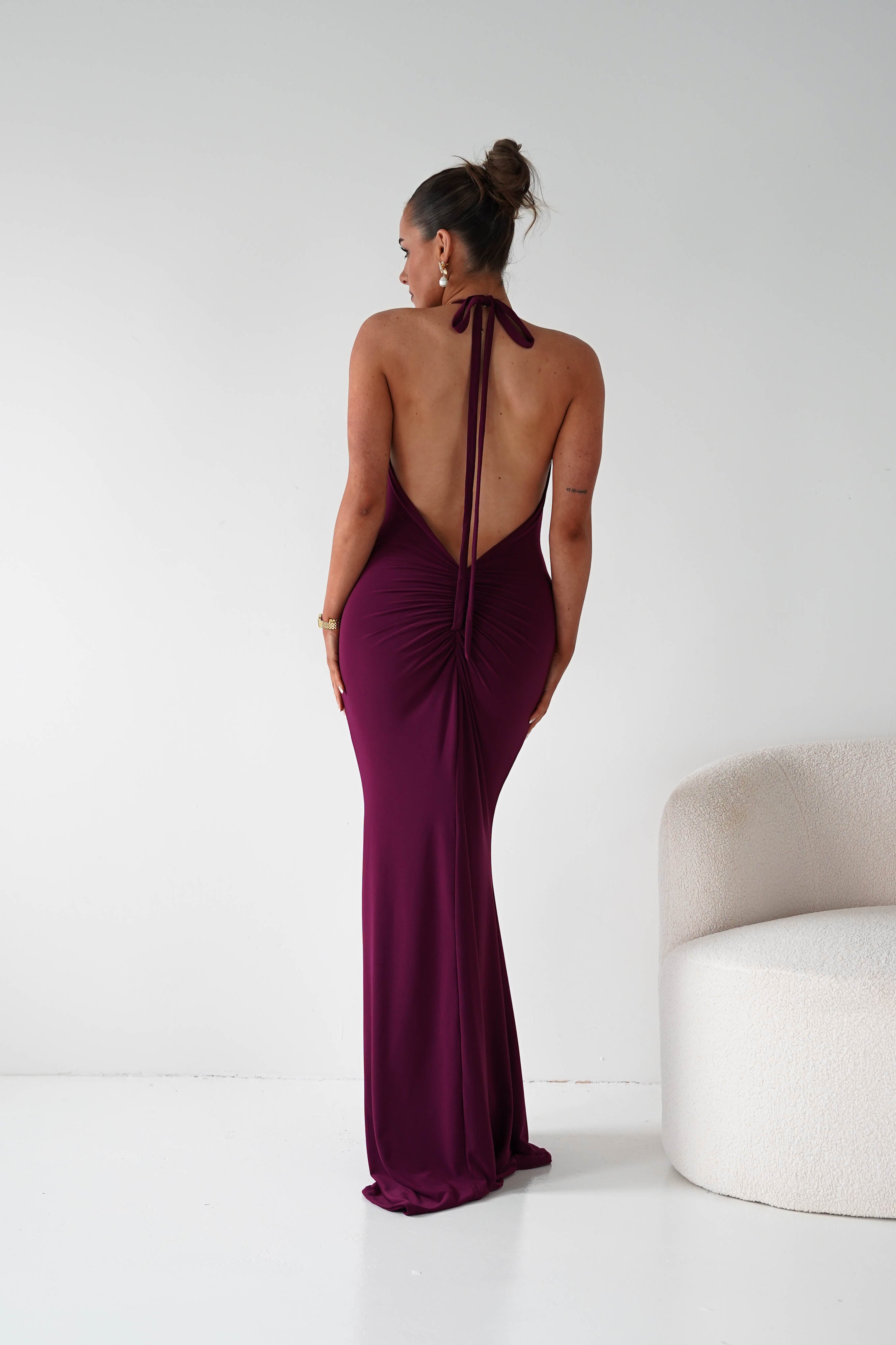 Woman wearing a burgundy evening gown with an open back in a minimalistic setting.