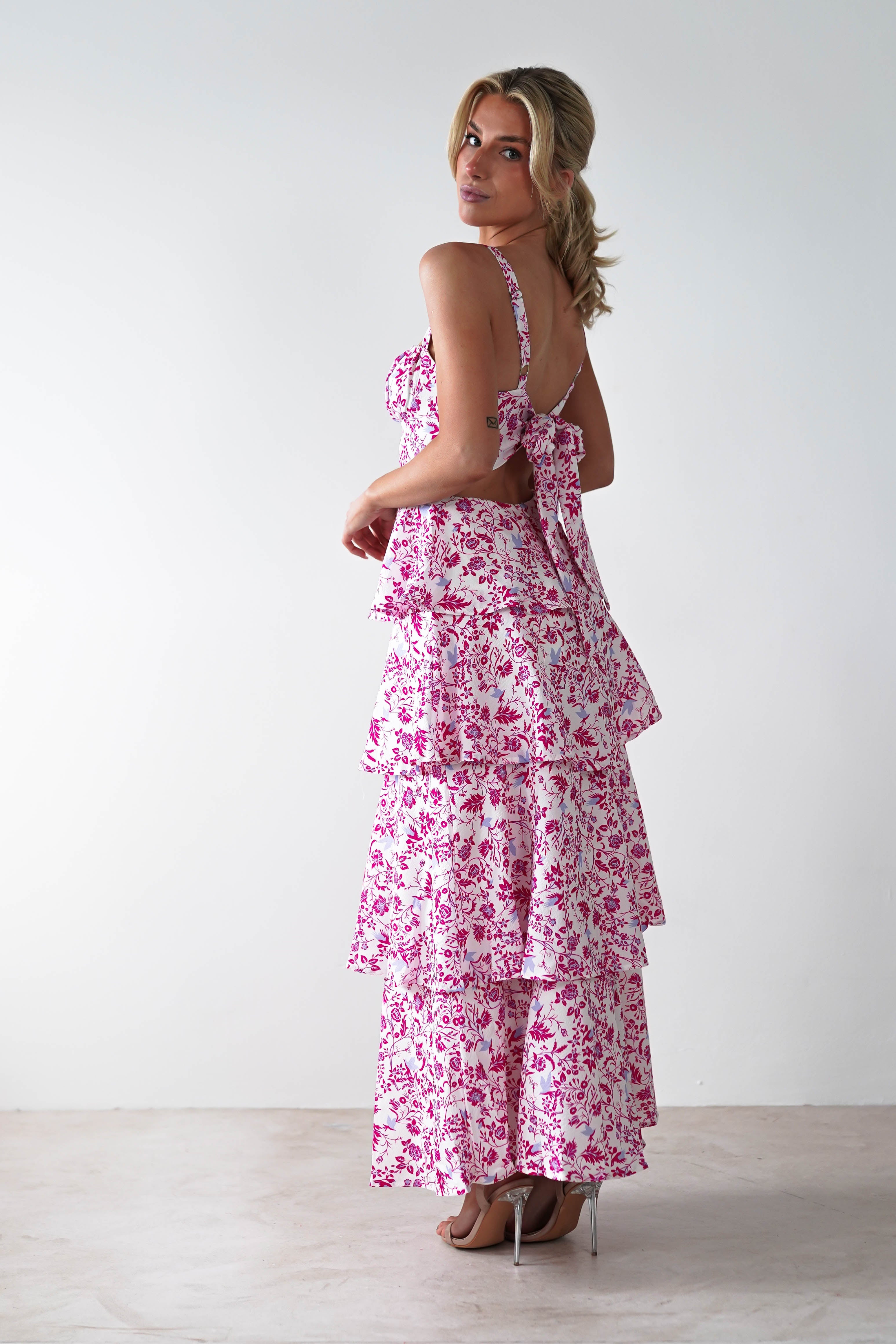 A woman in a ruffled floral pink and white summer print maxi dress stands against a plain white background. She looks over her shoulder, her hair in a loose ponytail. Perfect for wedding guests, the dress features layers, tie straps, and she pairs it with high heels.