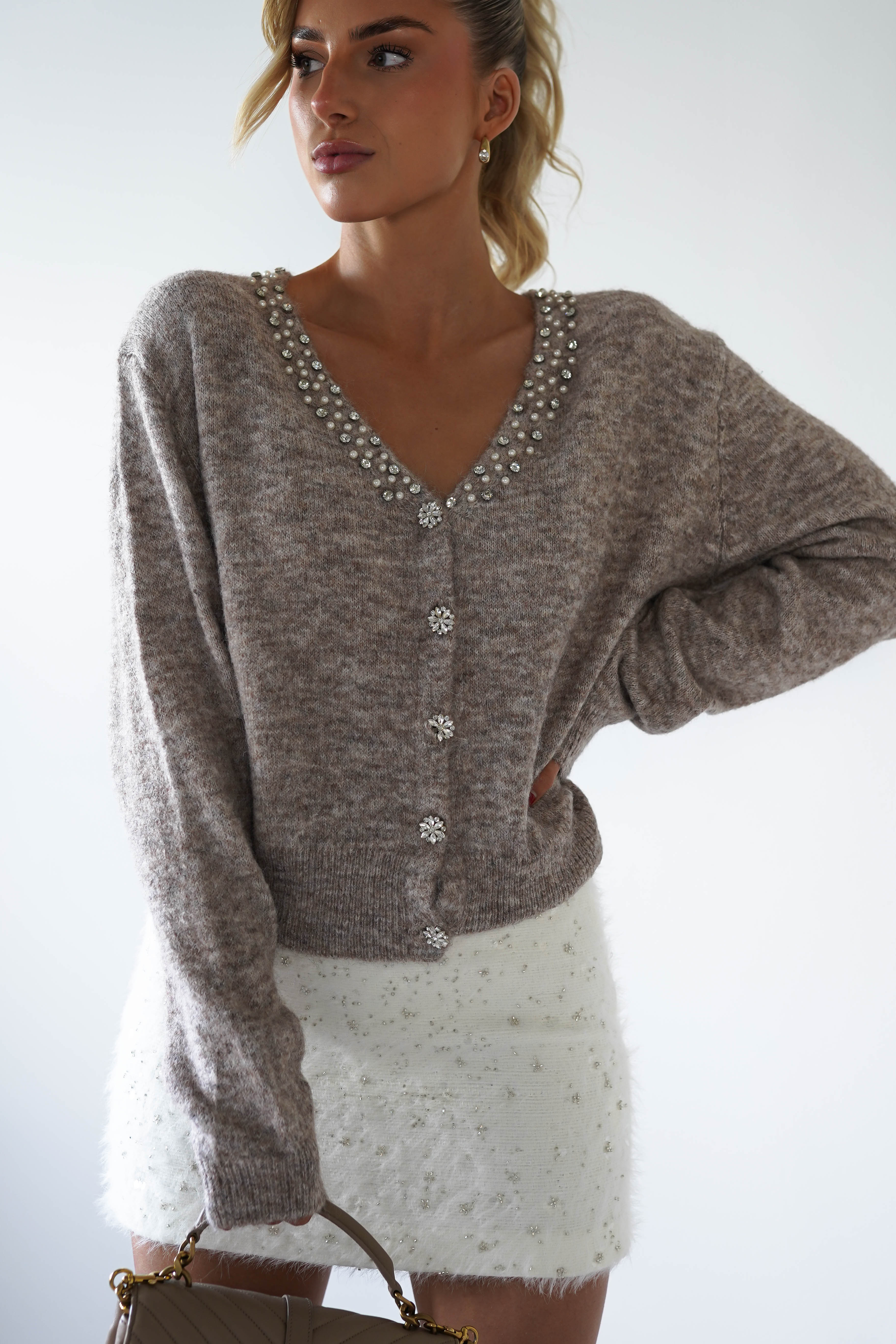 A person stands confidently, wearing a knitted cardi in textured beige with faux pearl detailing along the neckline and jewel buttons. Paired with a white skirt, they have their hand on their hip and exude poise against a plain white background.