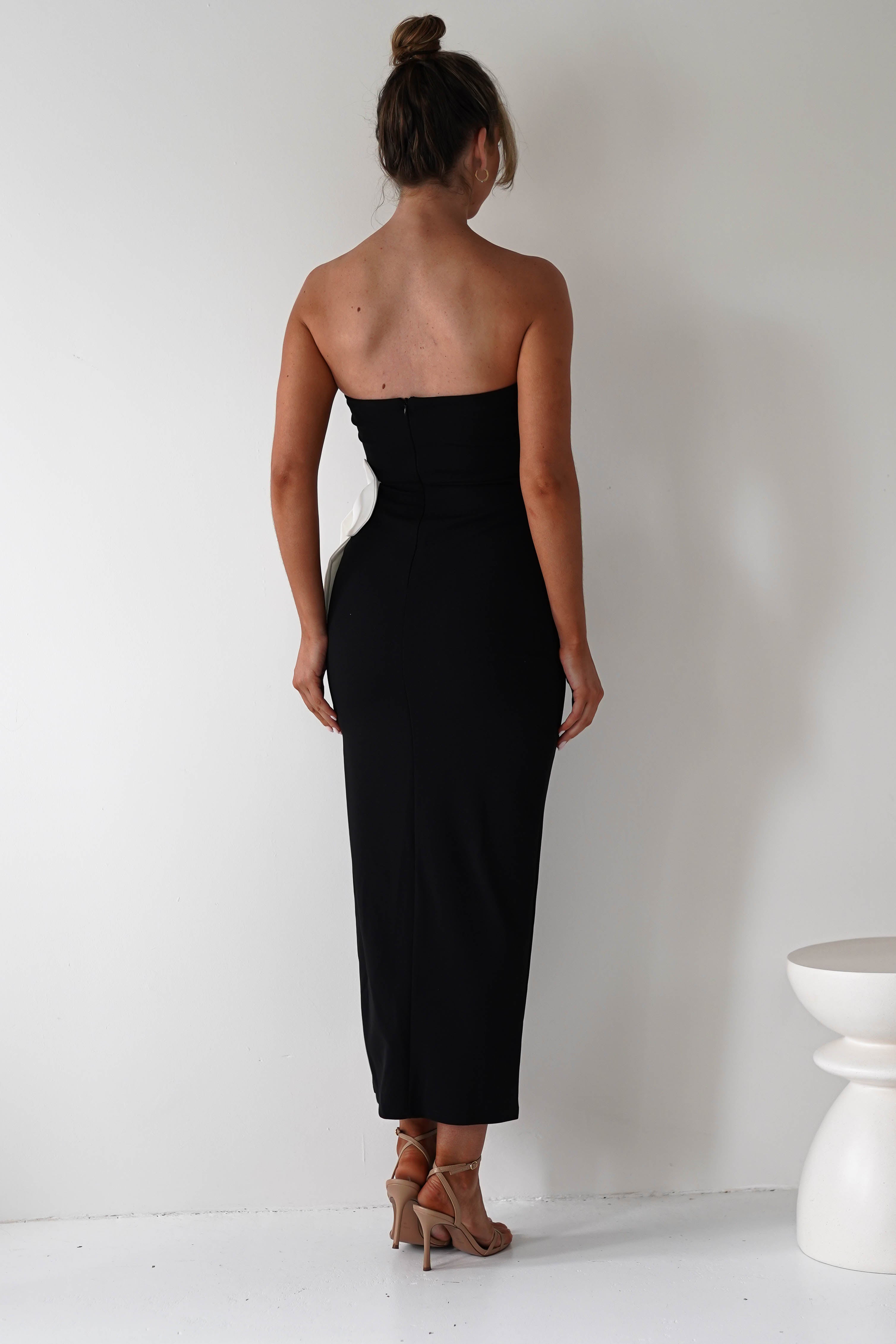 A woman in a strapless black midi dress and heels stands with her back to the camera. She is in a minimalist white room with a small white stool on her right. Her hair is in a bun, perfectly styled for an elegant wedding guest outfit.