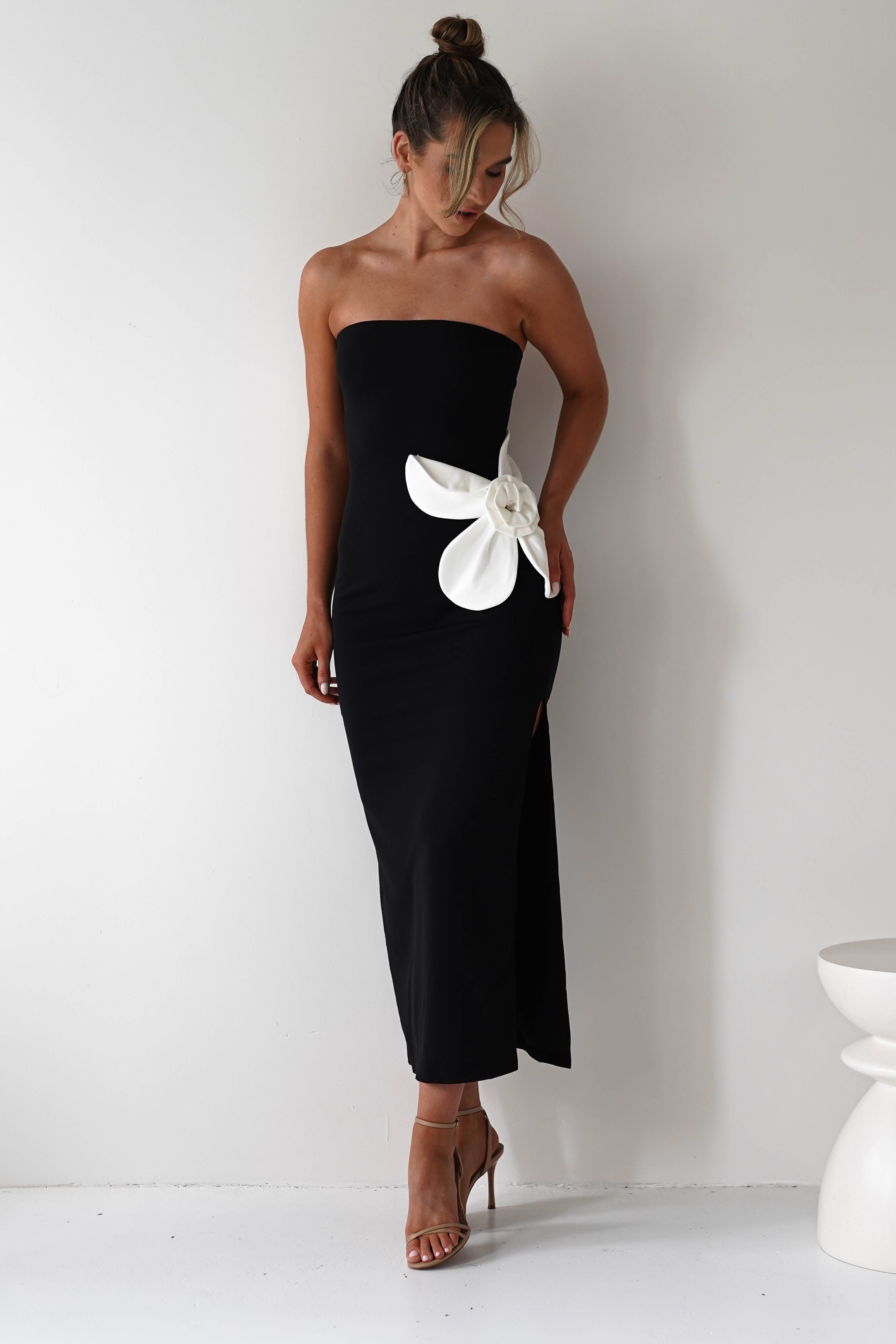 A woman in a strapless black midi dress with a striking floral feature on the side poses against a white background. Her hair is styled in a bun, and she complements the look with nude heels, making it an elegant wedding guest outfit.