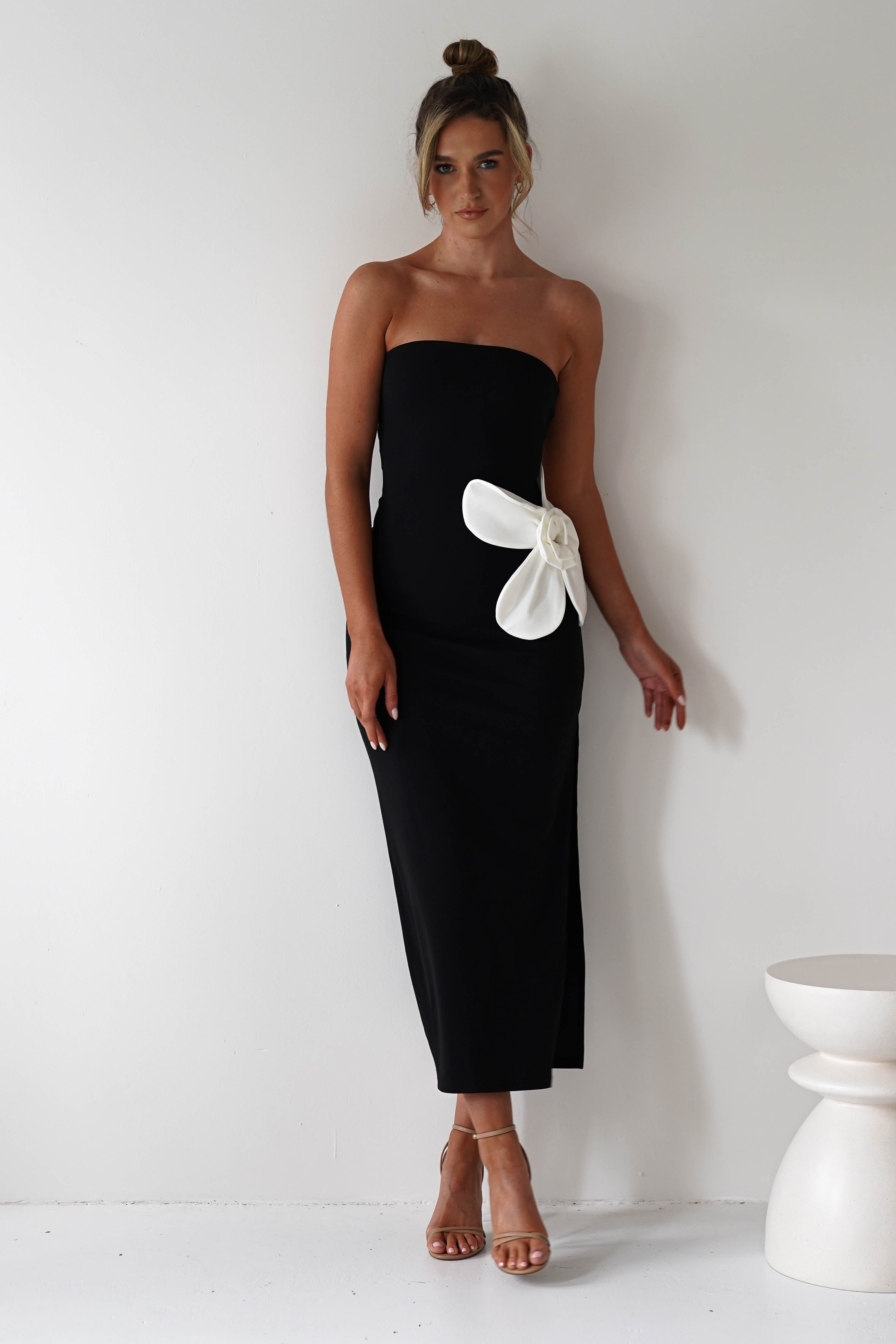 A woman in a strapless black midi dress with a striking floral feature stands against a white wall. Her hair is styled in an updo, and she dons strappy high heels. A white, minimalist stool sits to the right, perfecting this chic wedding guest outfit.