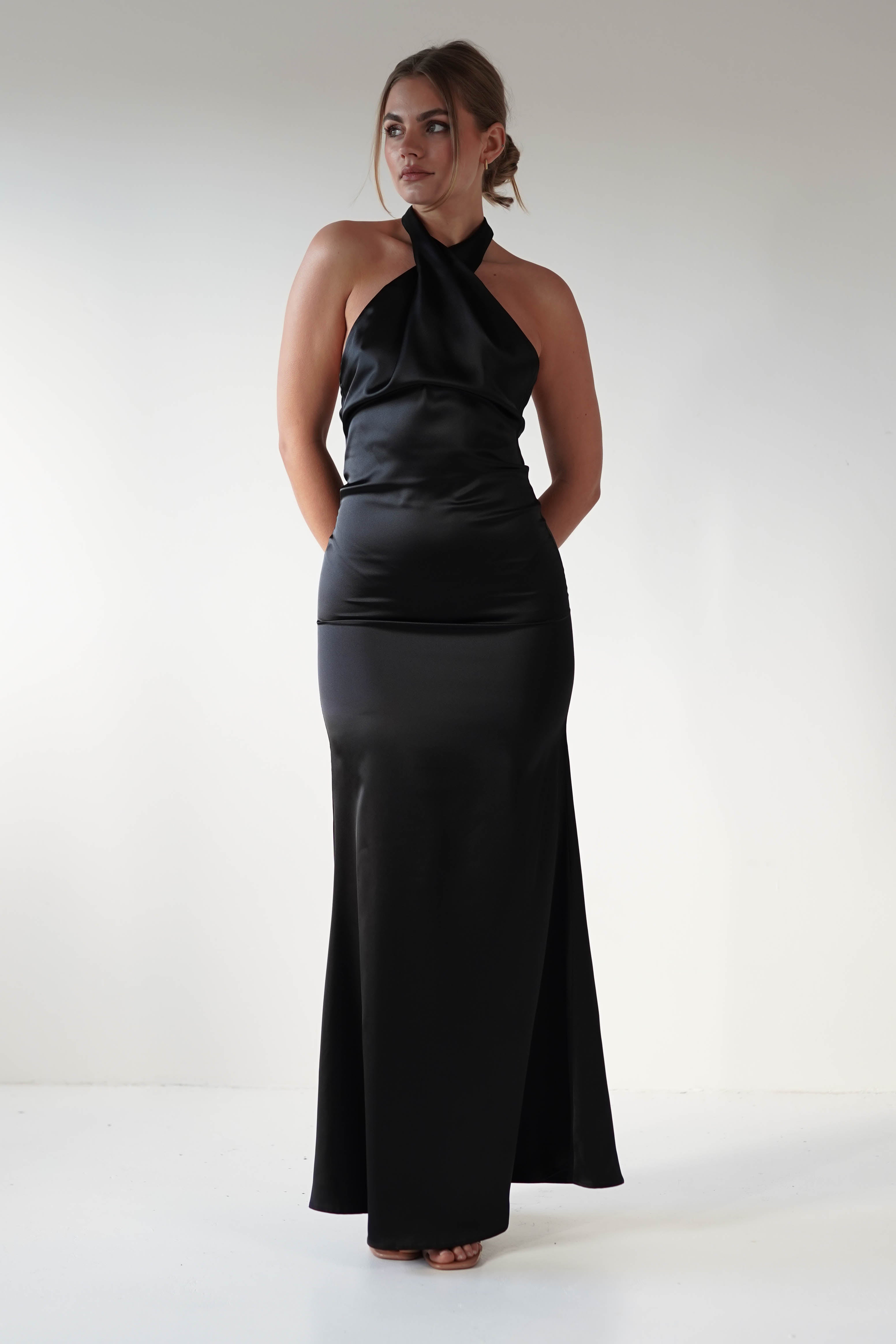 A person stands confidently in a sleek, sleeveless halter neck dress made of soft satin material. The open back design adds allure as the fitted silhouette elegantly flows to the floor. Against a plain white wall, their hair is styled up, enhancing the sophisticated look.