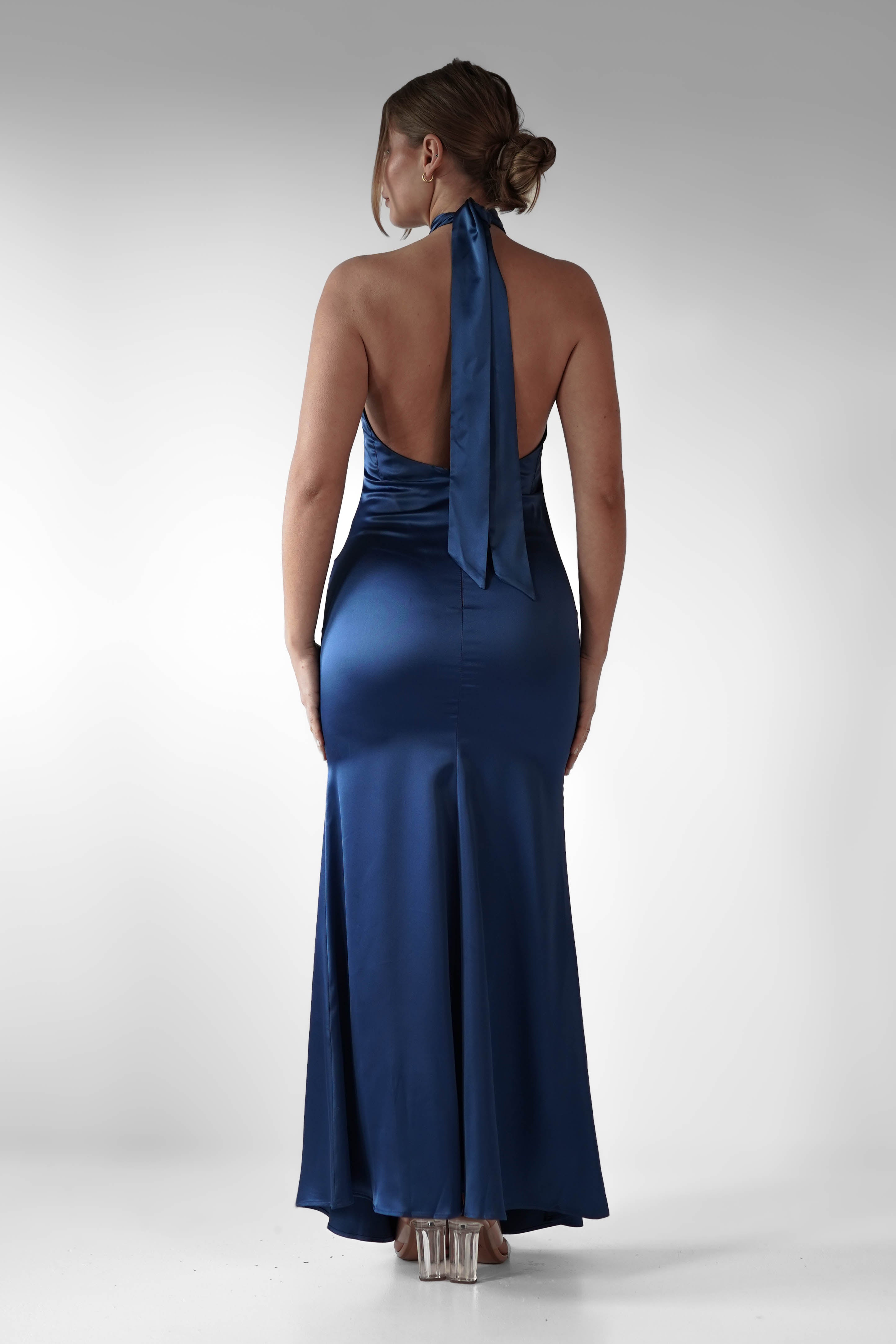 A woman in a halter-neck, backless blue satin gown stands facing away, embodying elegant wedding guest attire. The dress cascades to the floor, complemented by her heels. Her hair is styled in an updo against a plain white background.