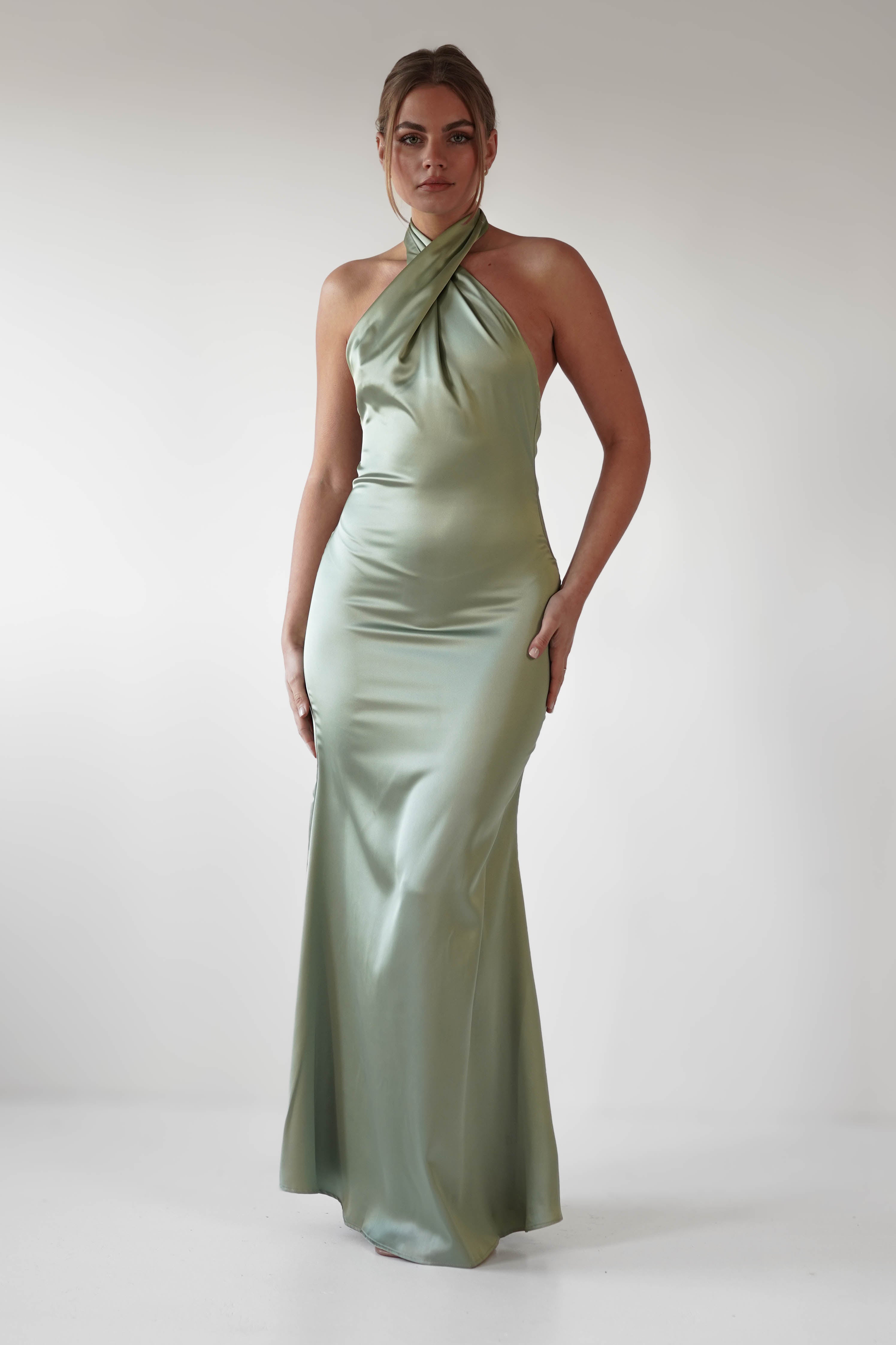 A woman wearing a long, silky green halter neck dress stands against a plain white background. The satin material of the form-fitting wedding guest attire drapes elegantly to the floor as she looks towards the camera with a neutral expression.