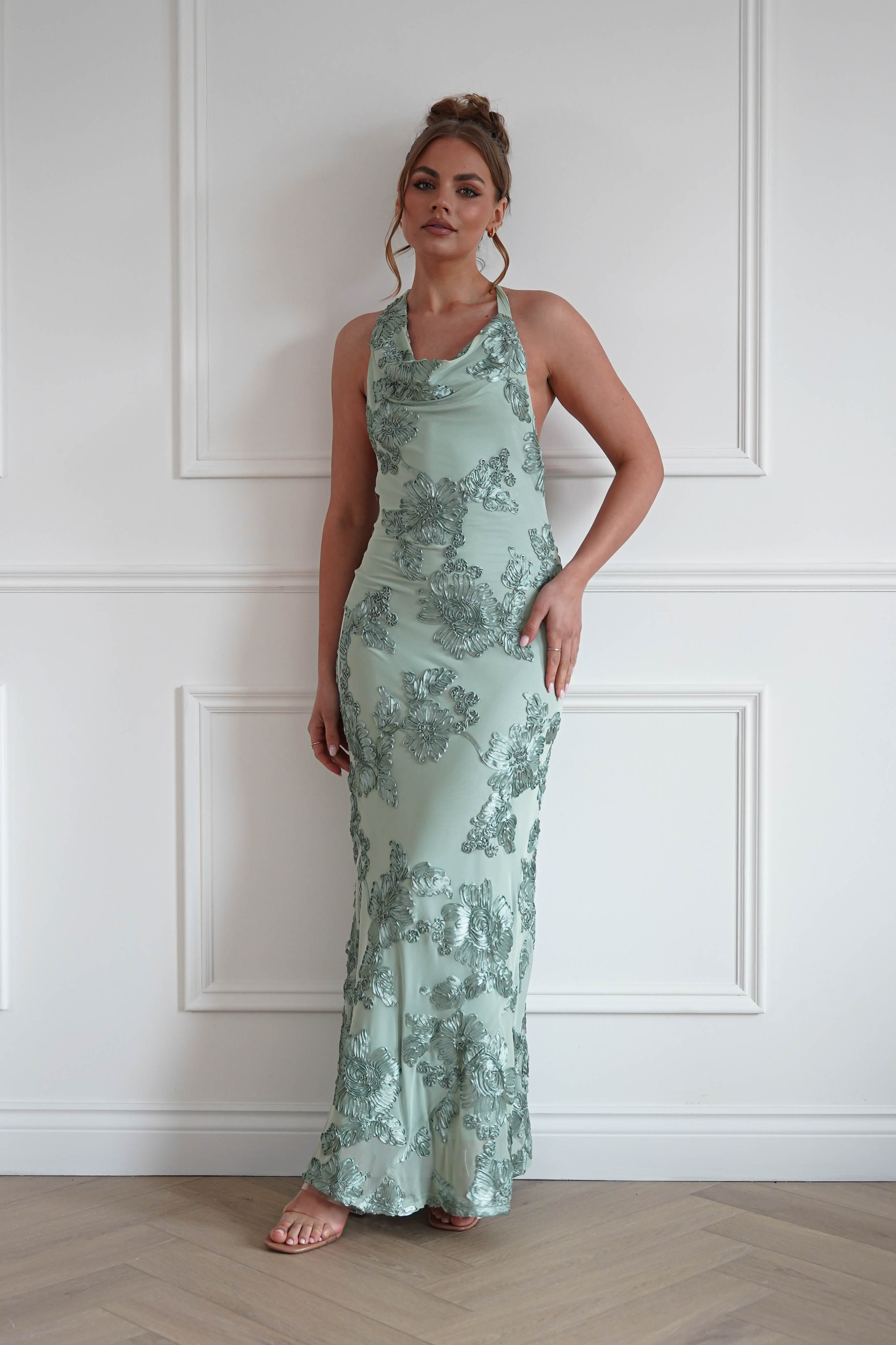 Woman wearing a sage green halterneck maxi dress with floral embroidered embellishments, a draped cowl neckline, and a fitted floor-length silhouette, perfect for weddings, race days, formal events, and special occasions.